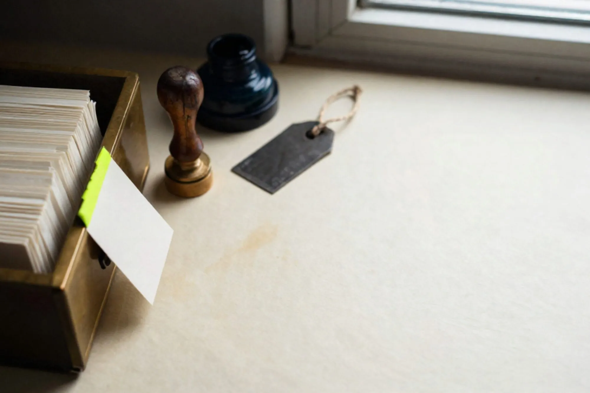 Open brass card-index drawer with cream cards, one chartreuse tab, wooden stamp, iron tag on ivory desk.