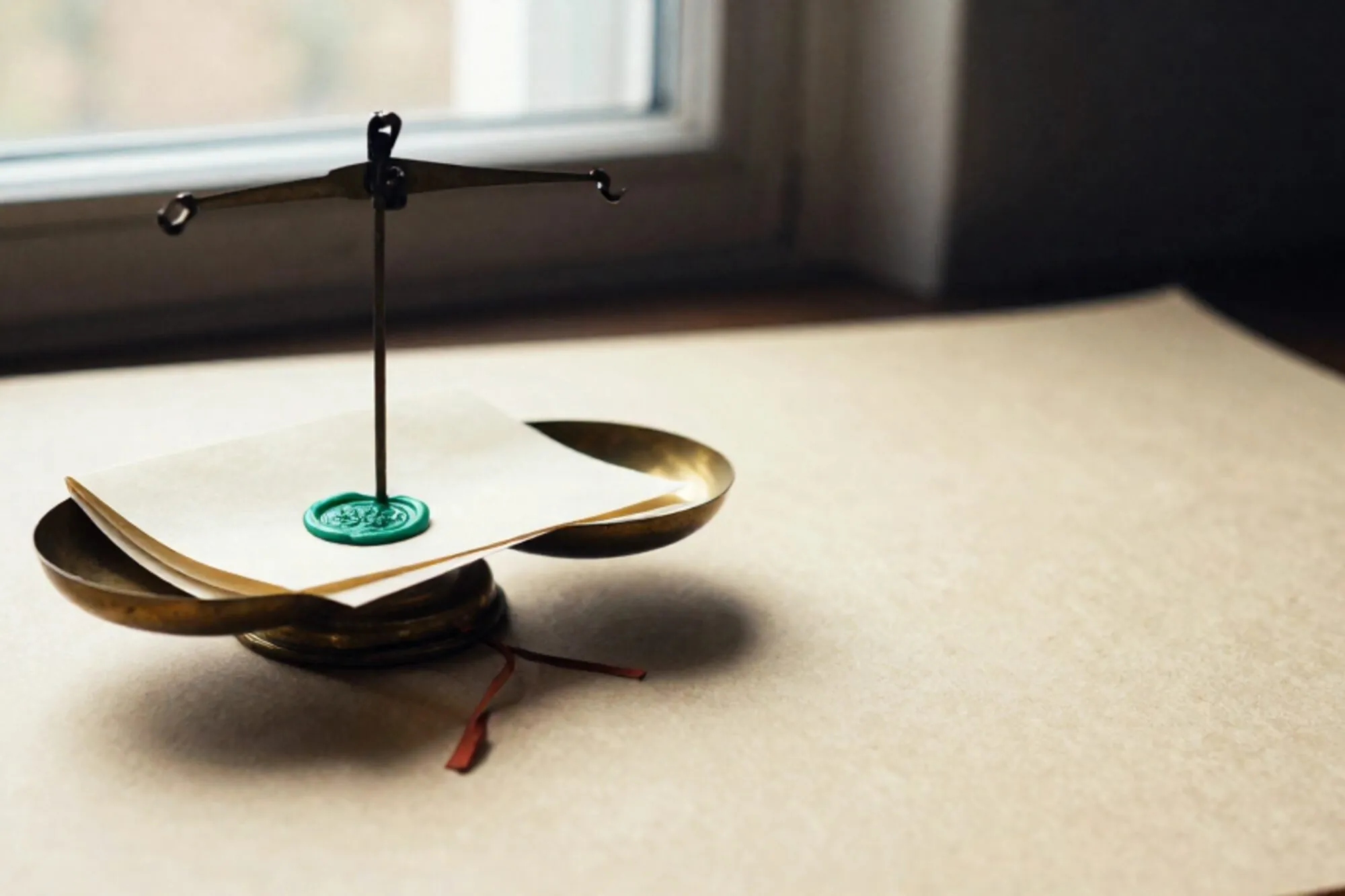 Worn brass weighing scale on ivory paper, two folded cream slips in pans, chartreuse wax seal, clay-red ribbon, soft shadows.