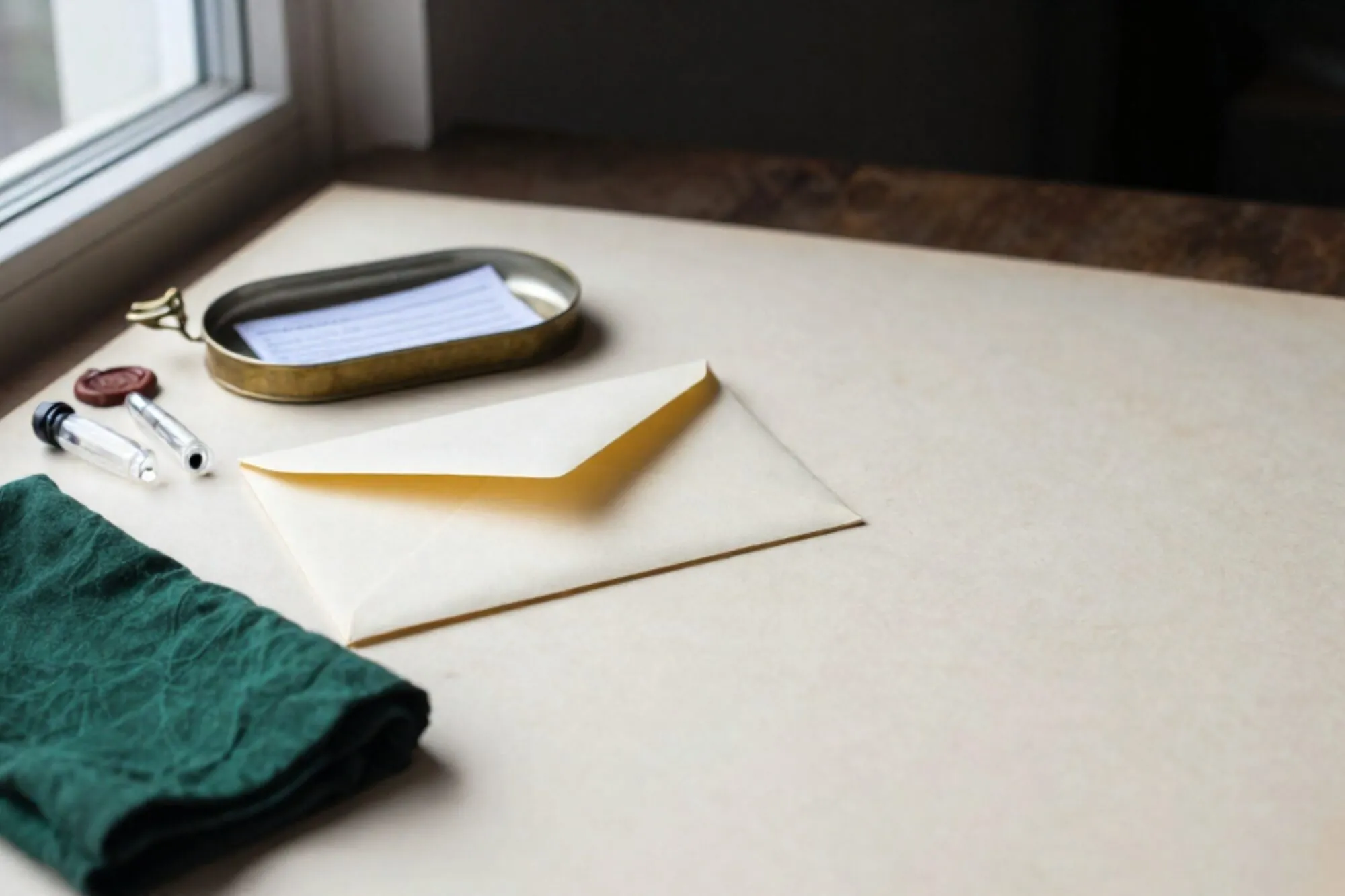 Crème envelop met groen lint, koperen schaaltje met glazen flesjes, papieren briefje onder klem, gevouwen groene doek.