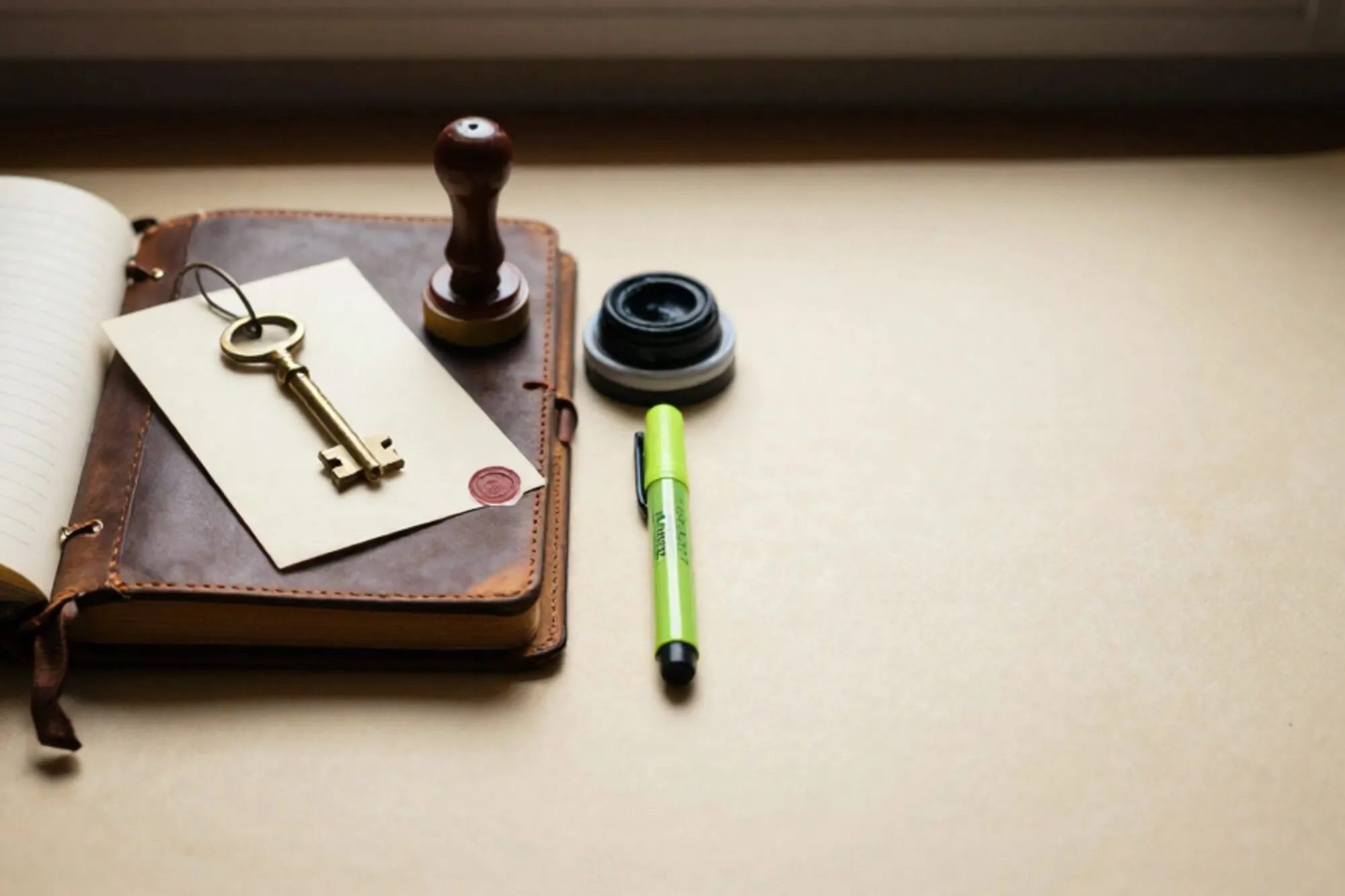 Open leather logbook, brass key on cream index card, rubber stamp, ink pad, green ribbon, red wax fragment on ivory desk.