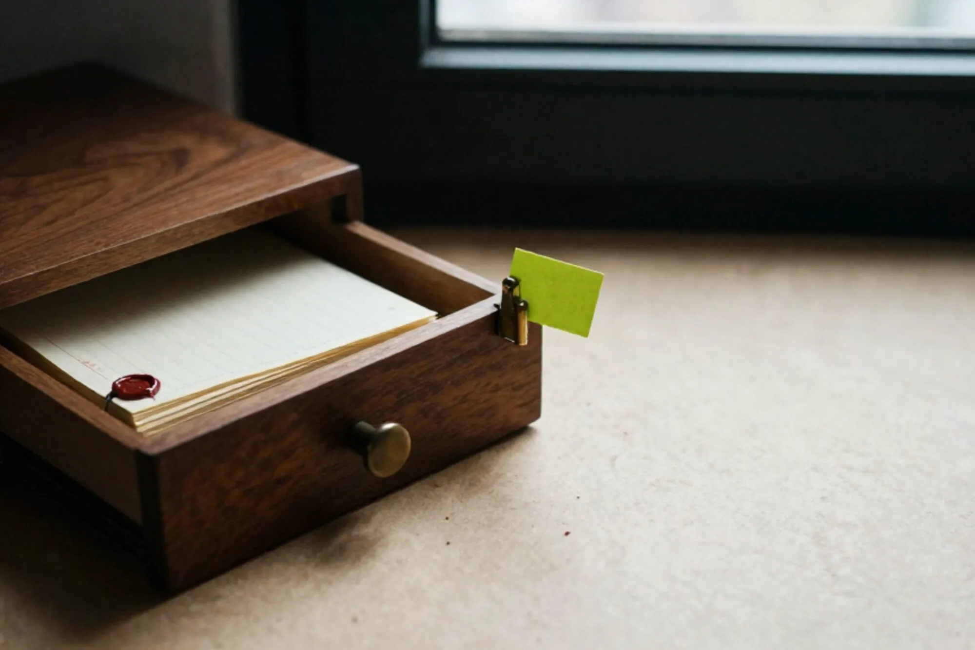 Open wooden index-card drawer on ivory desk, brass divider with chartreuse tag, ledger paper stack, red wax seal.