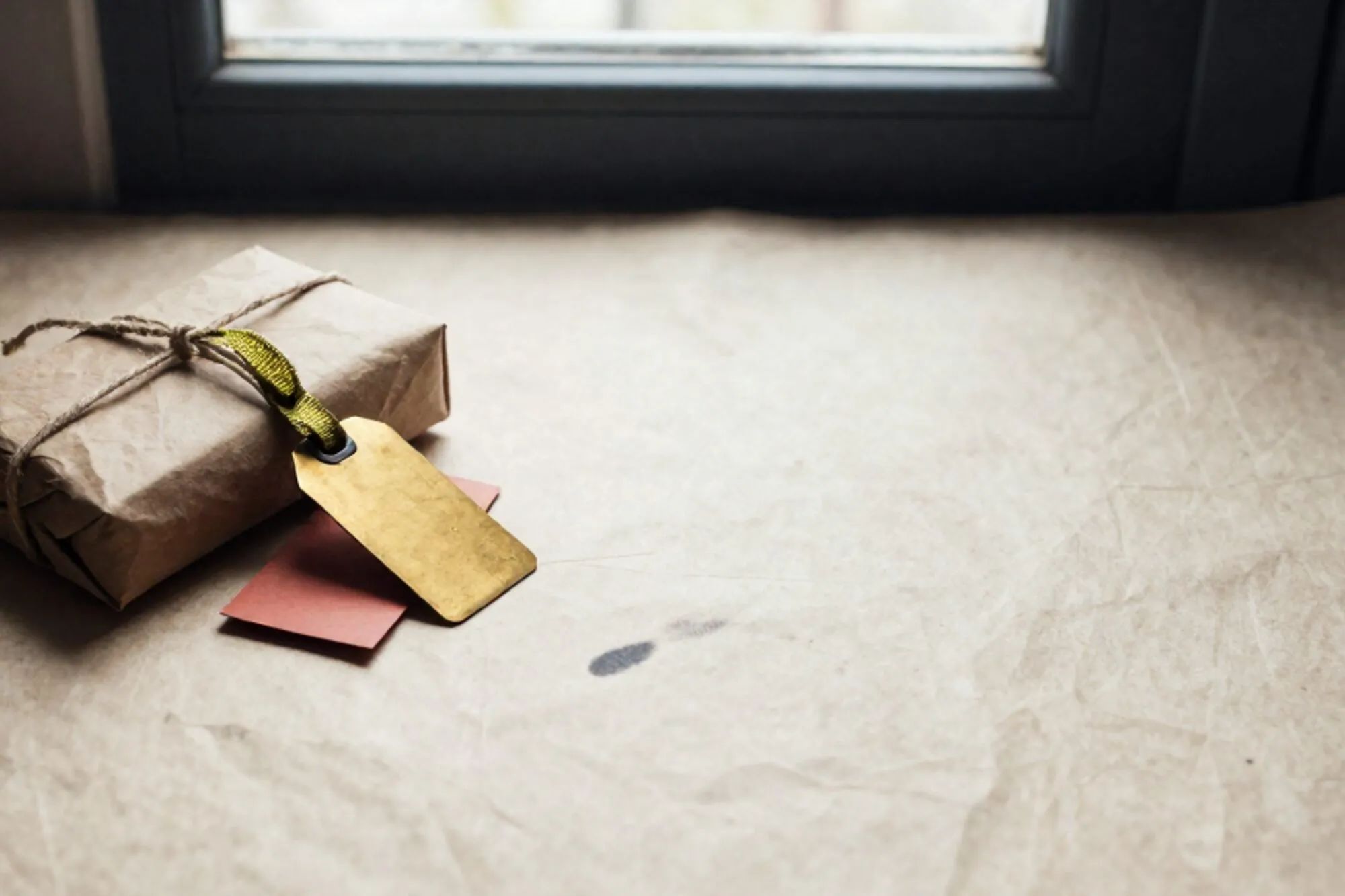 Small brown parcel with linen twine, brass luggage tag with green ribbon, red shipping label on ivory linen desk.