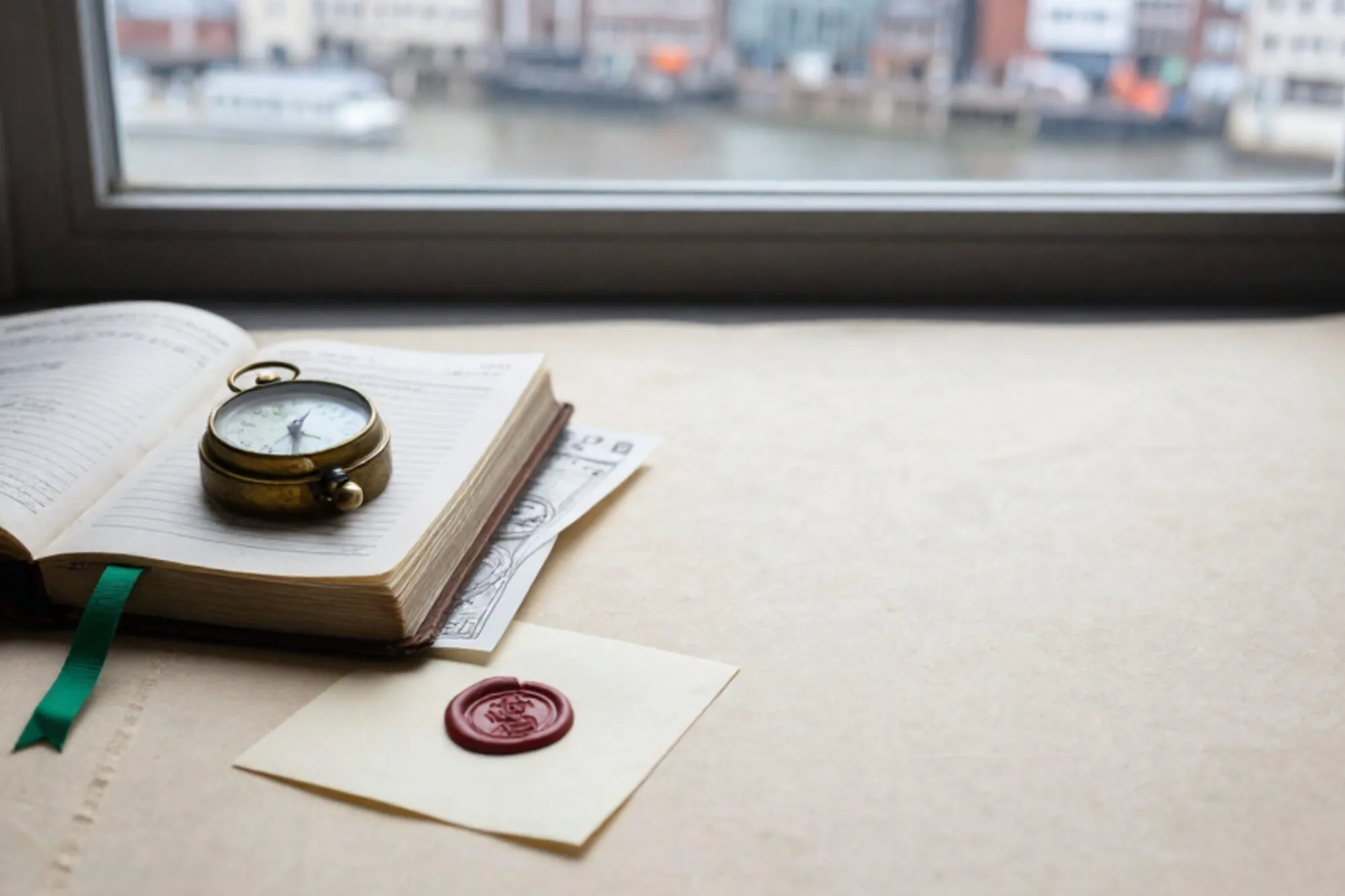 Open leather ledger with brass tide dial, green ribbon and red wax seal on a cream linen desk by a port window.