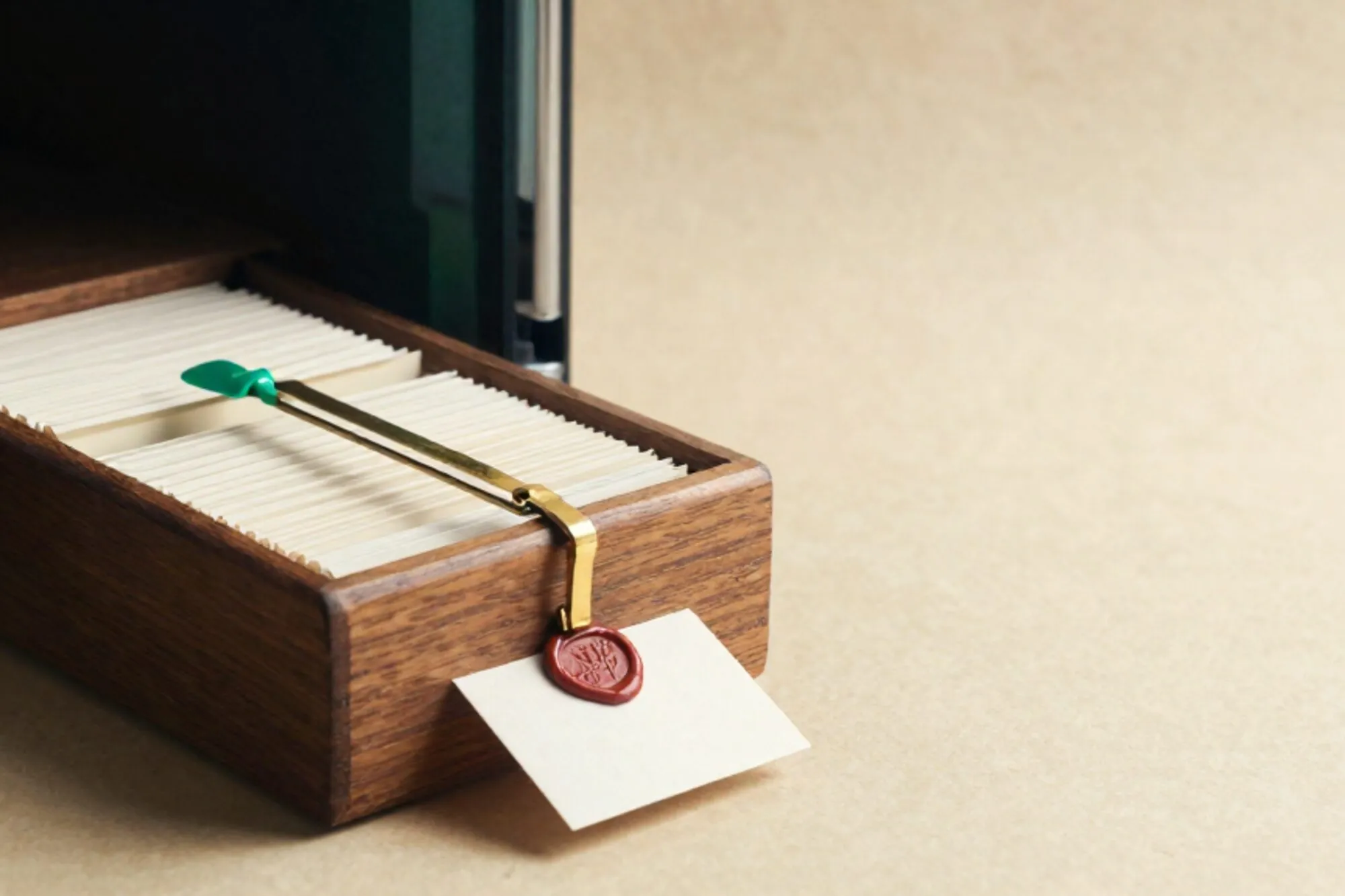 Wooden index-card drawer open at a green tab between cream cards, brass divider, wax-sealed card, ivory background.