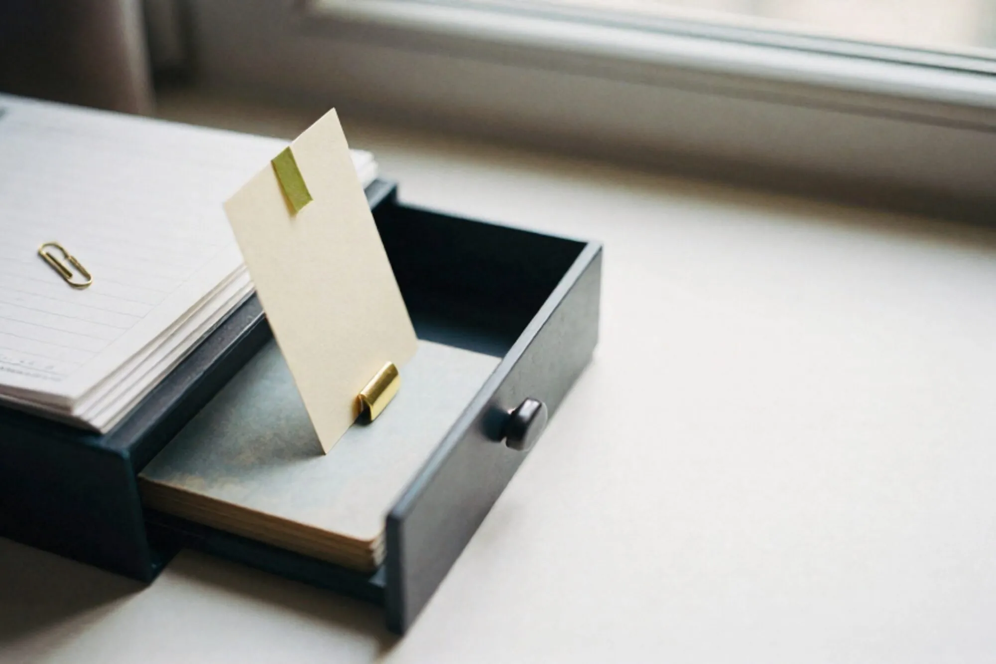 Open oak index-card drawer on a pale desk, one cream card lifted on a brass tab with a green ribbon, ledger paper beside it.