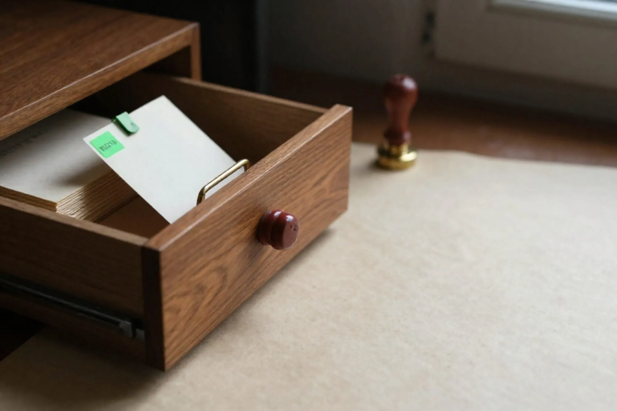 Open oak index-card drawer on ivory desk, one card raised on a brass tab with a green clip, red wax seal beside.