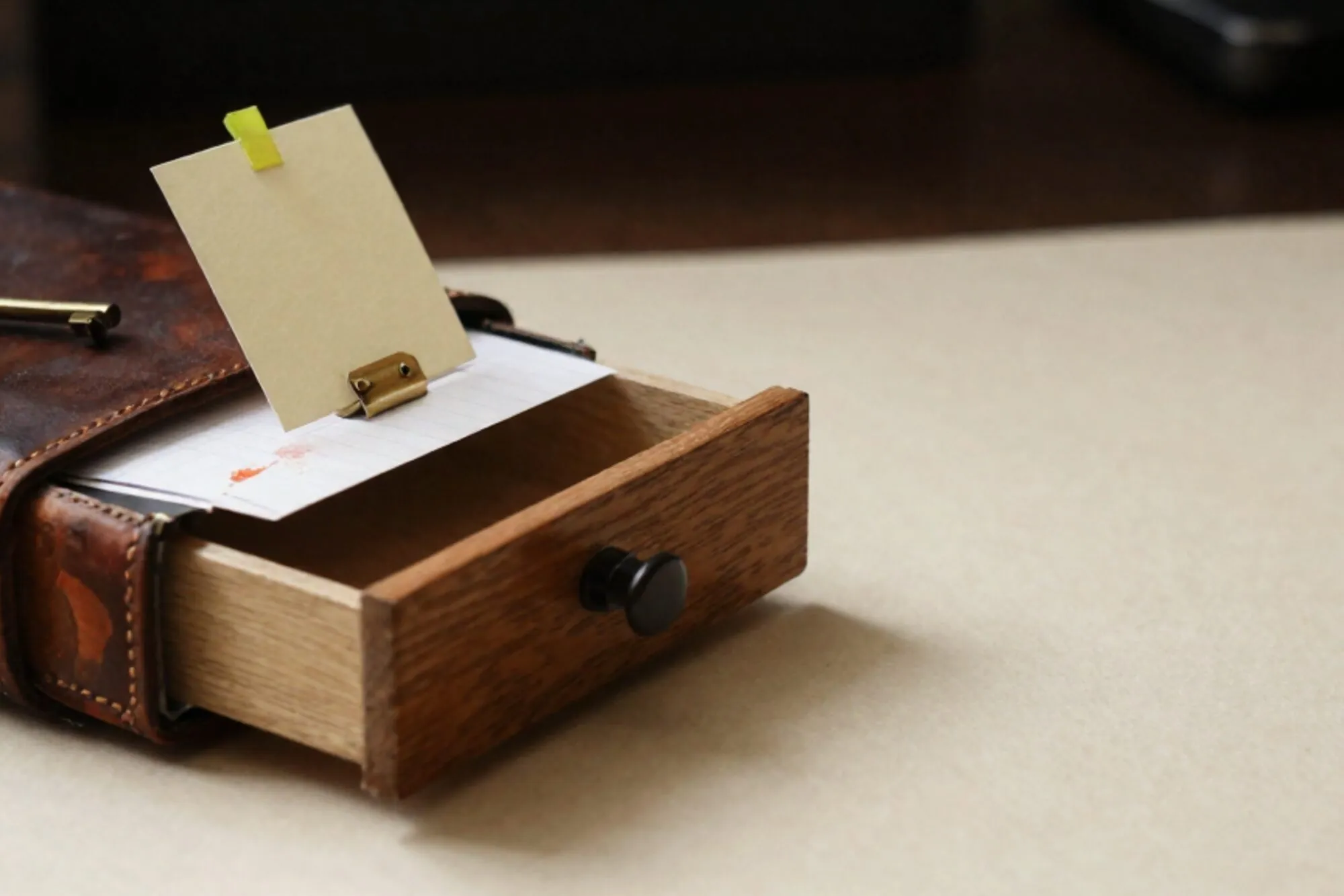 Open oak index-card drawer on ivory desk, one cream card raised by brass tab with green ribbon, leather logbook and brass key beside it.