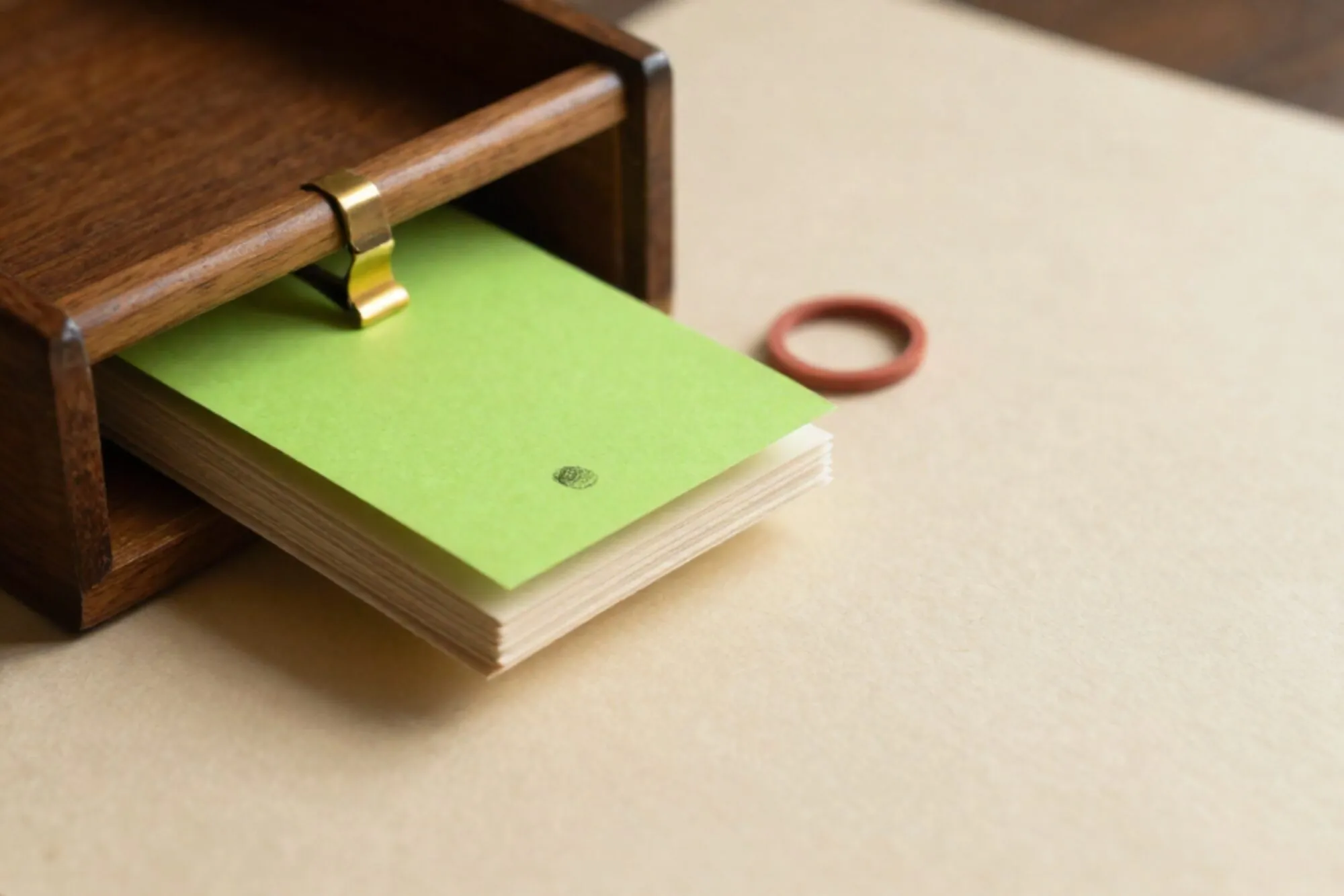Open wooden index-card drawer on ivory paper, one green card raised, brass divider, red rubber band beside it.