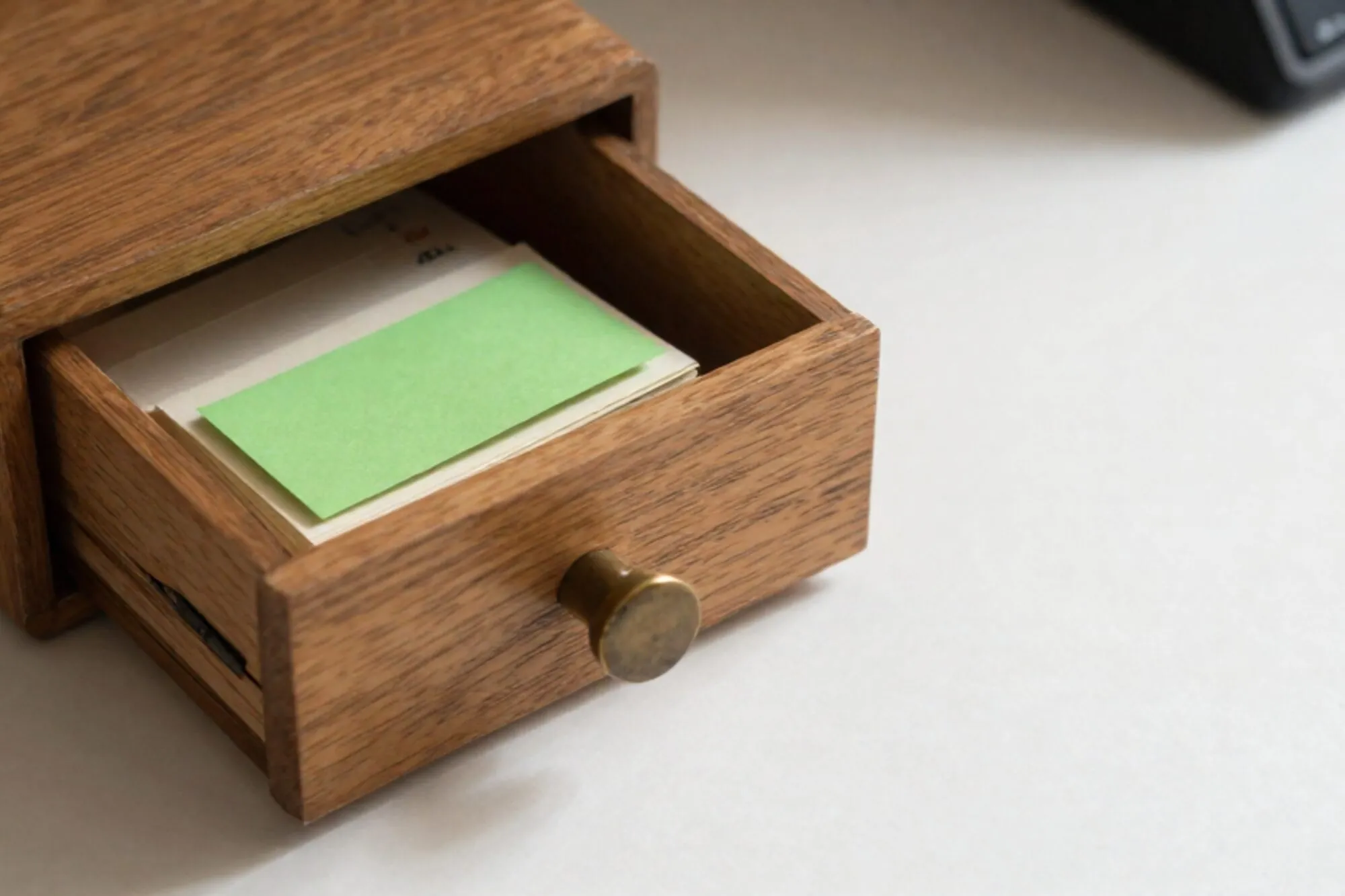 Open oak index-card drawer on pale desk, brass divider askew, one green card among cream cards, soft side light.