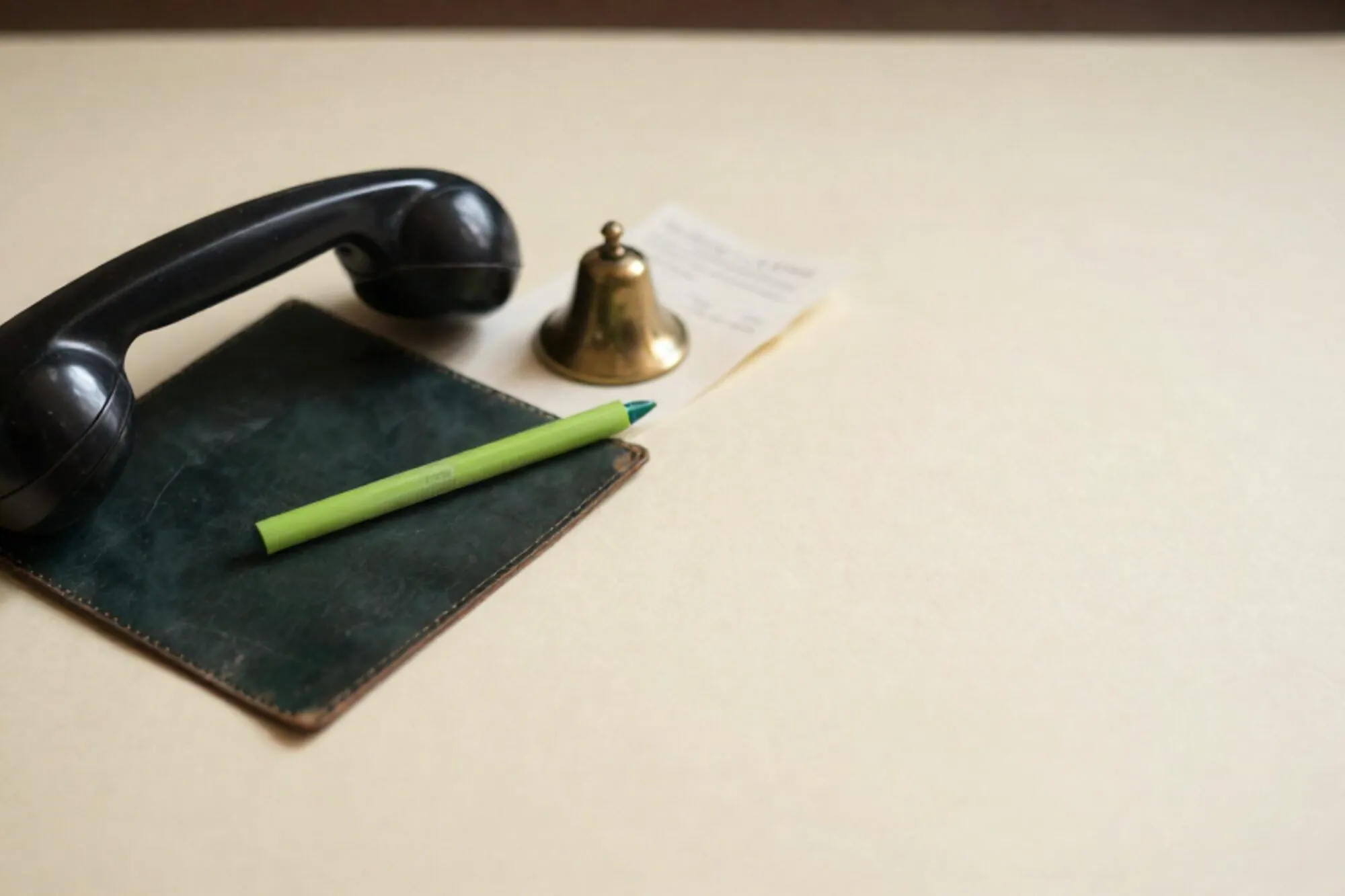 Zwarte bakelieten telefoonhoorn op donkergroene leren onderlegger, limoenlint, koperen bel, gevouwen papier op ivoor.