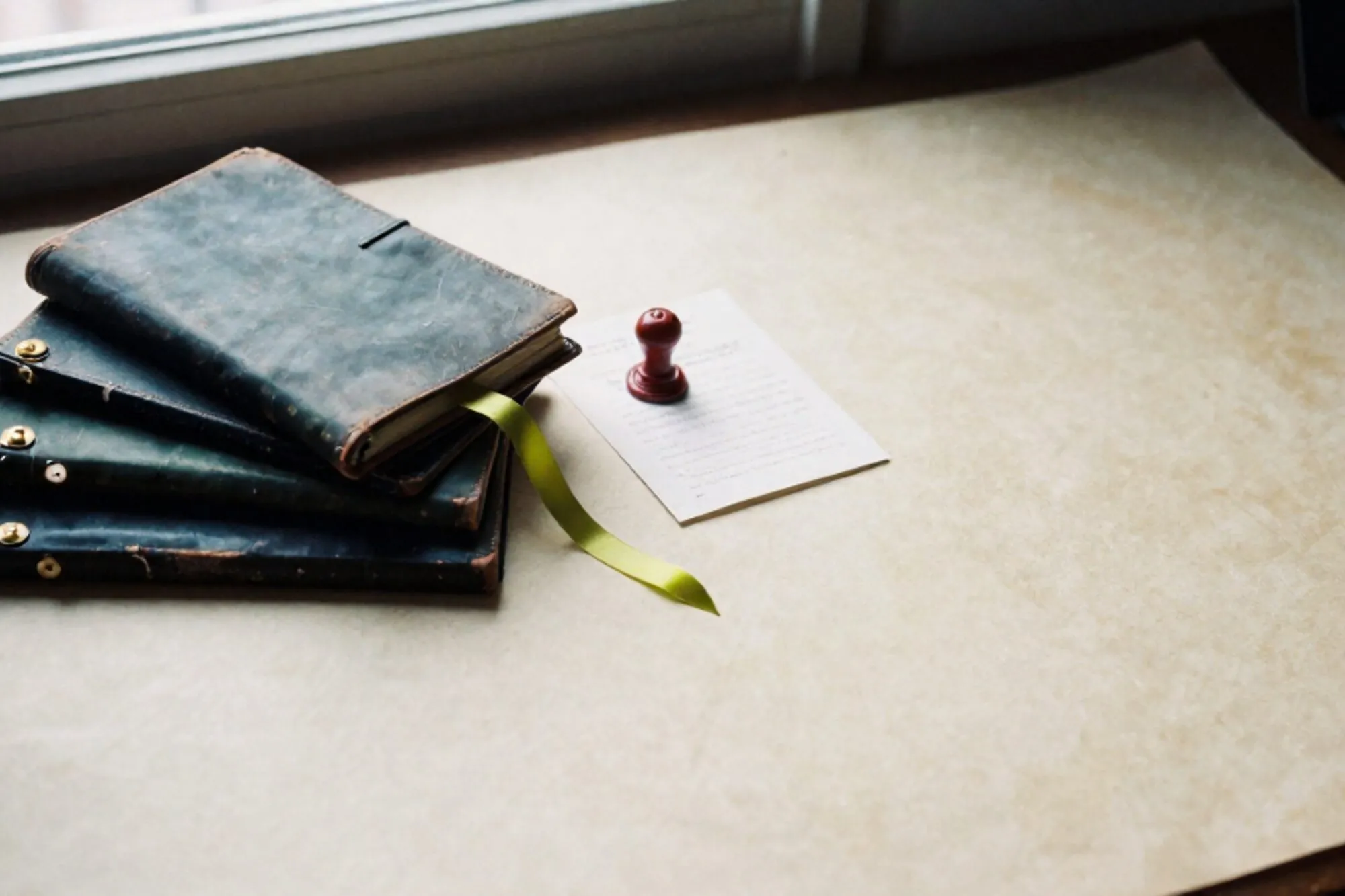 Three stacked leather notebooks with brass tabs, green silk ribbon, red wax seal on card, ivory paper desk.