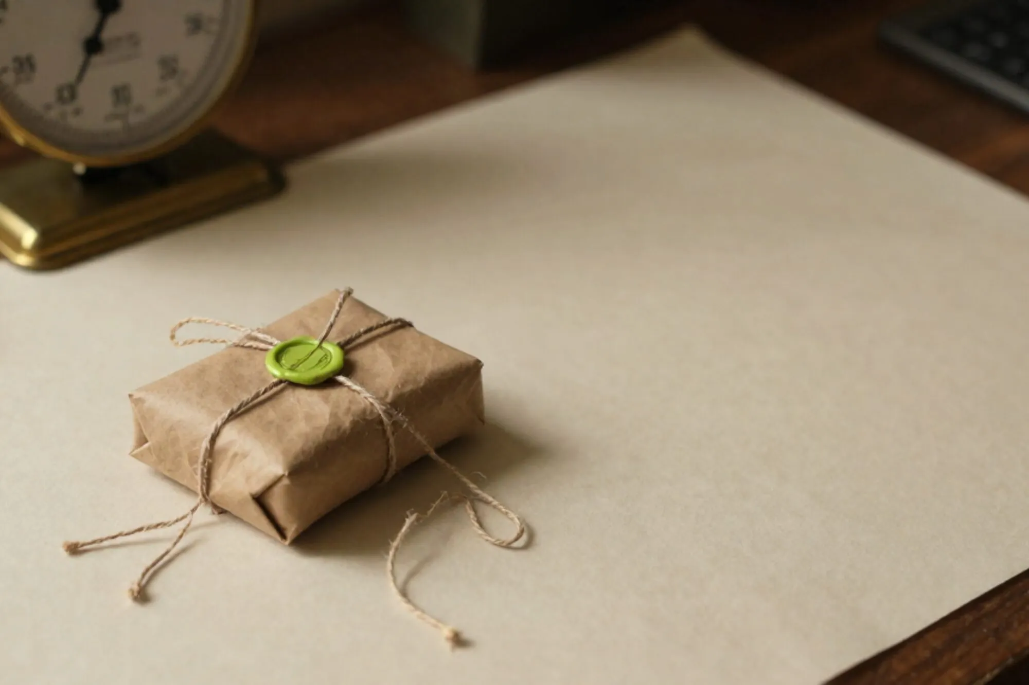 Half-unwrapped craft parcel with seven twine knots, brass postal scale, green wax seal, red stamp on ivory desk.
