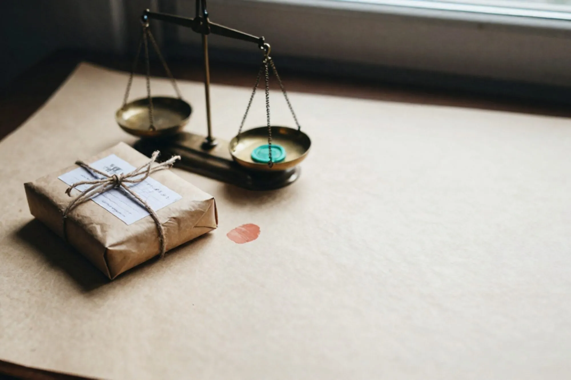Small craft-paper parcel with frayed twine beside a tilted brass scale holding a green wax seal on ivory desk.