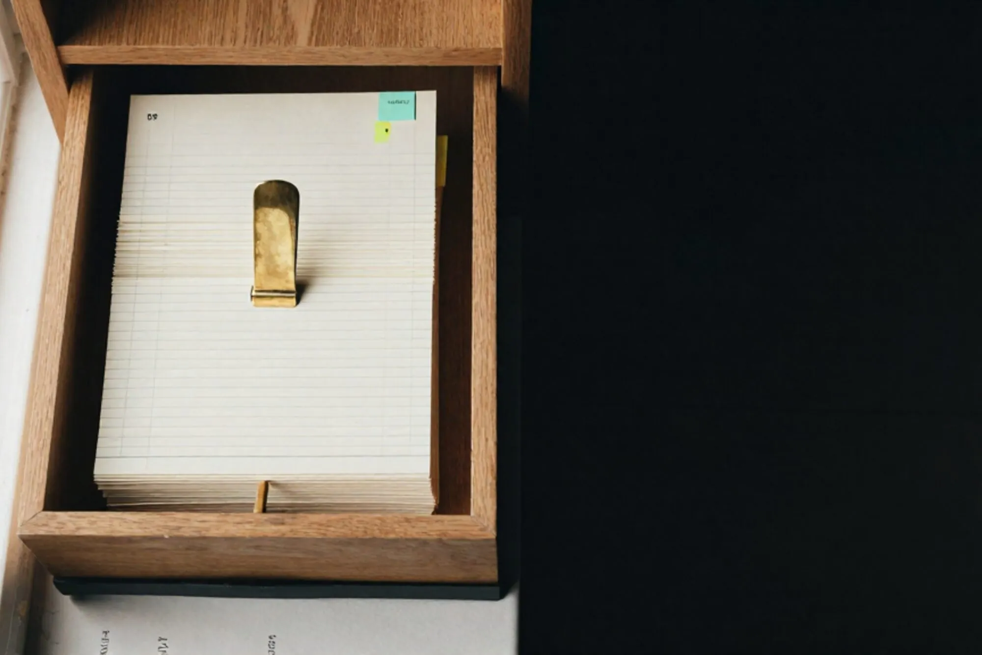 Open oak index-card drawer with cream cards, brass divider, green tab, ledger bundle on ivory paper.