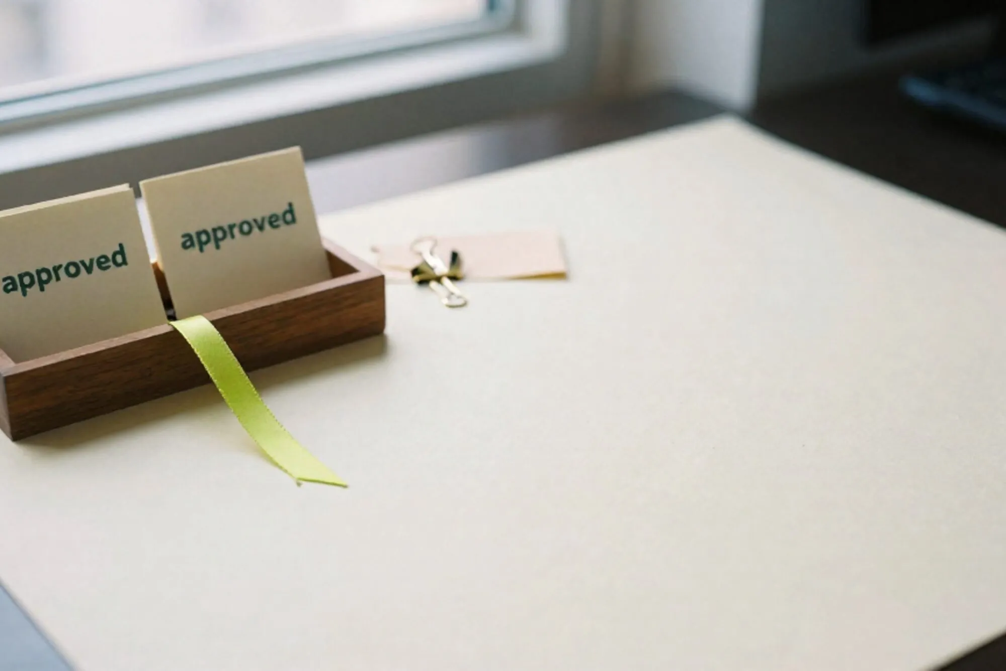 Wooden card tray with two cream index cards stamped approved, green ribbon, brass paper clip, manila slip on ivory desk.