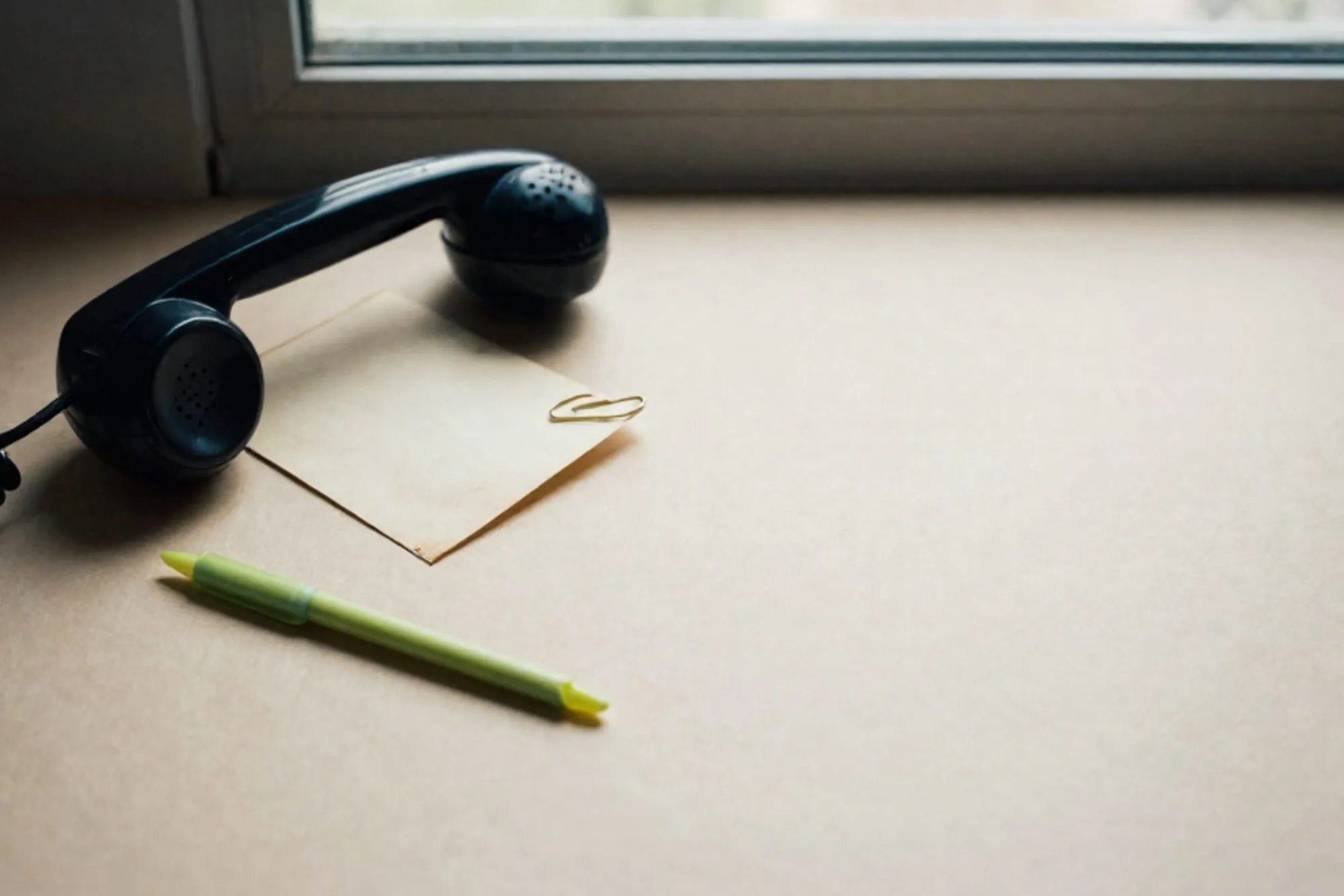 Black bakelite phone receiver off-hook on ivory blotter, coiled cord, green ribbon, cream index card with brass clip.