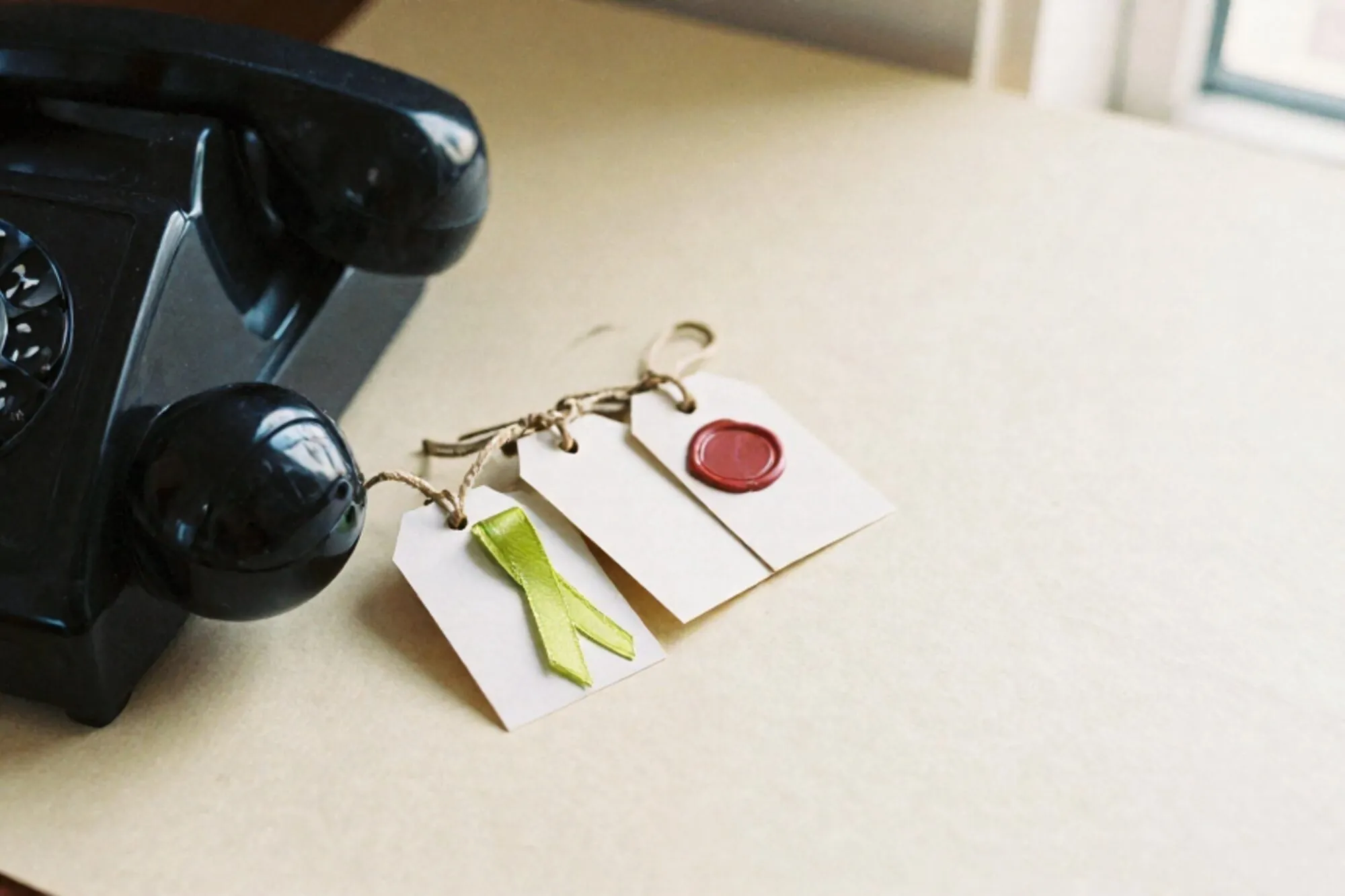 Black bakelite phone receiver off-hook on ivory paper, three twine-tied paper tags beside it, one with green ribbon, one with red wax seal.