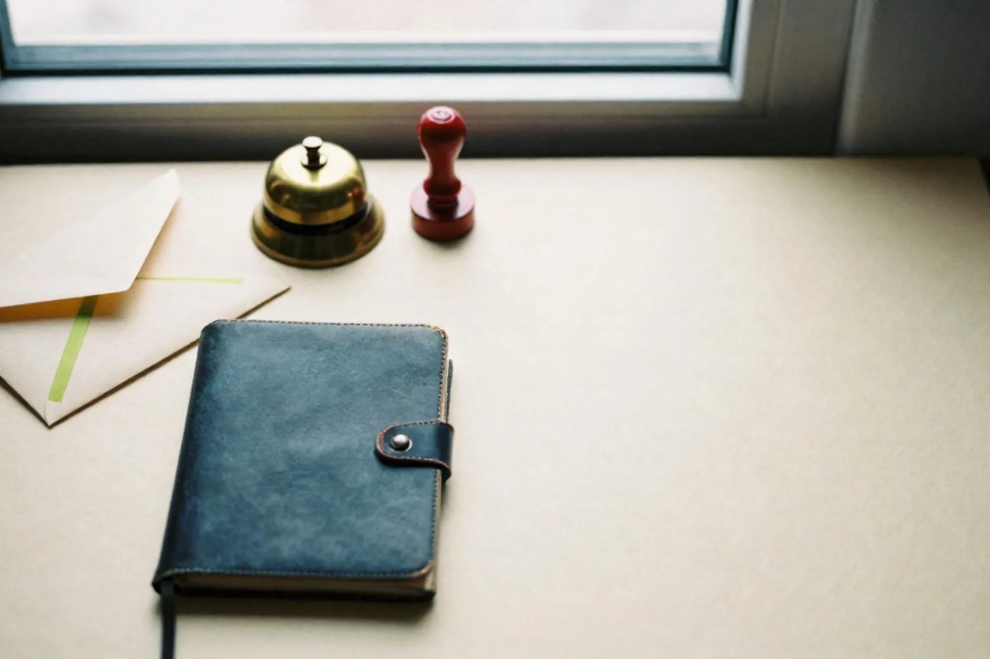 Cream envelope with green ribbon, brass bell, leather ledger and red wax stamp on ivory desk by window.