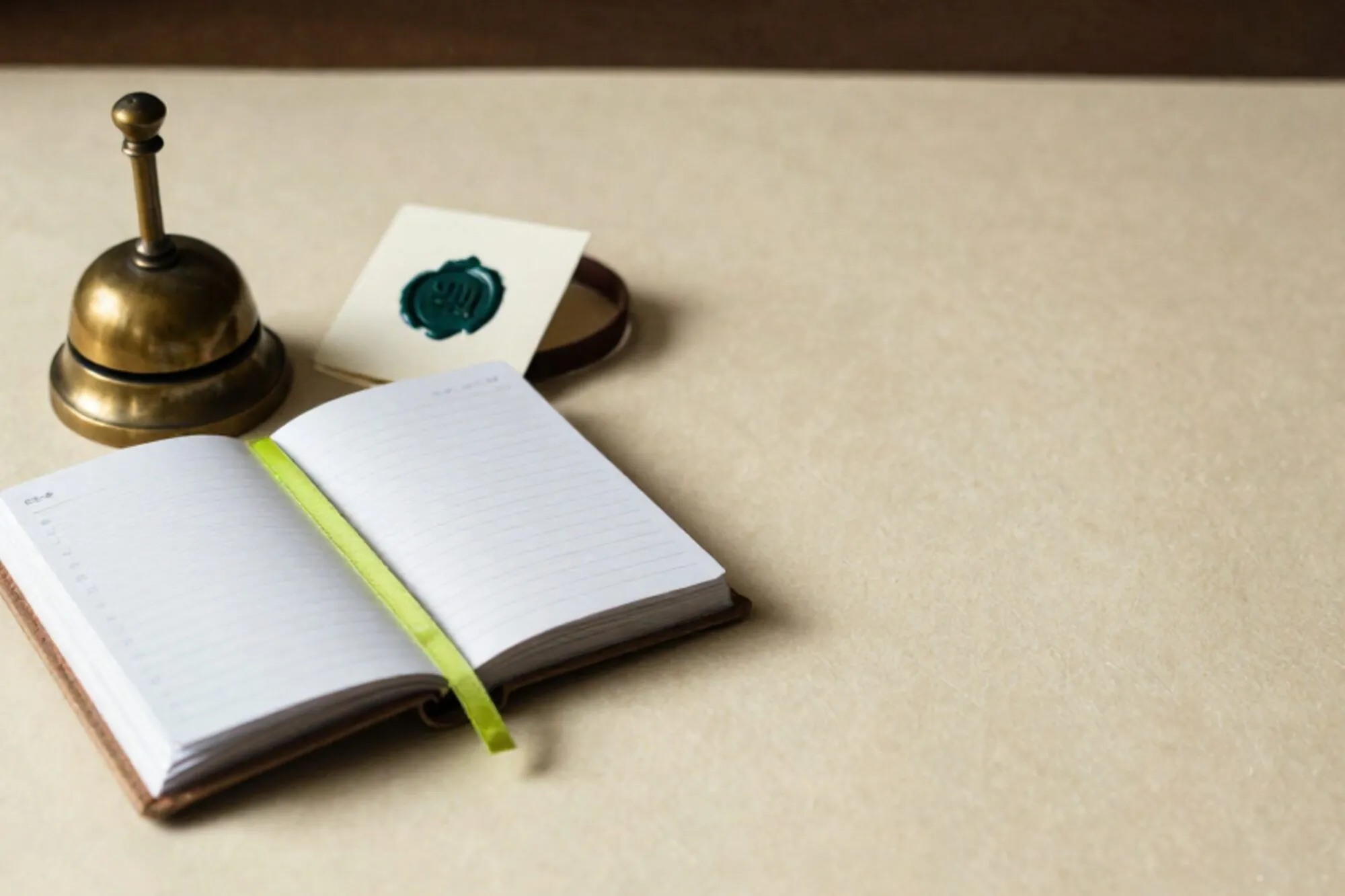 Linen ledger open with chartreuse ribbon, brass bell, wax-sealed card, leather collar on ivory paper.