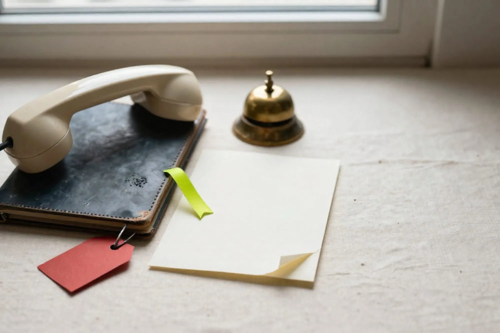 Crème bakelieten telefoon van haak op donkergroen leren vloeiblad, gekrulde snoer, notitieblok met groen lint, koperen bel, rood label.
