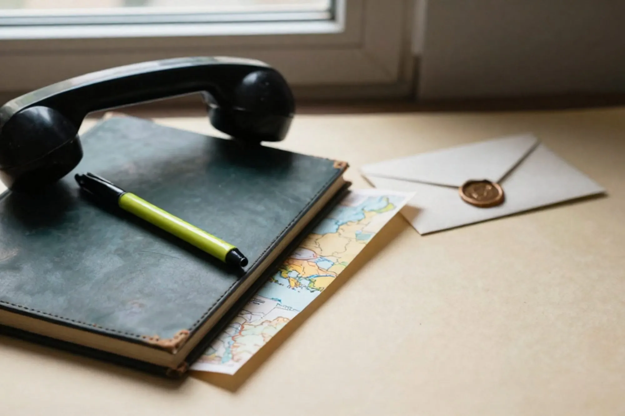 Black bakelite phone receiver off-hook on dark green leather blotter, green ribbon, folded paper map, wax-sealed envelope, brass weight.