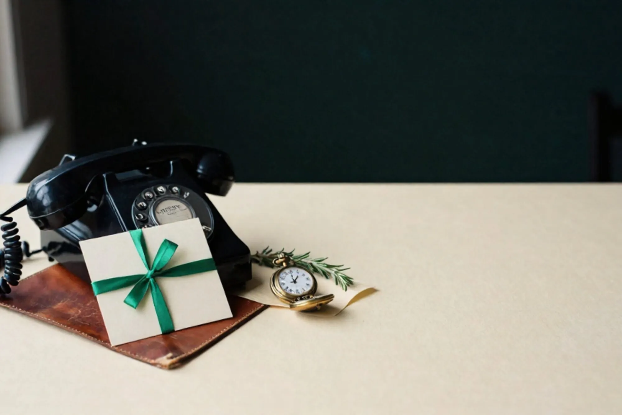 Black bakelite phone receiver off-hook on leather blotter, ivory card with green ribbon, brass pocket watch, dried rosemary.