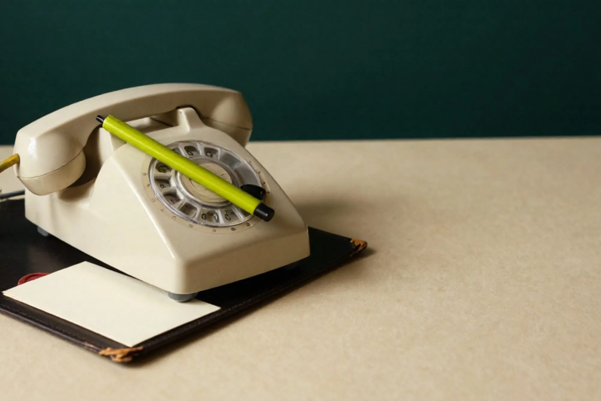 Cream bakelite phone receiver off-hook on dark leather blotter, green ribbon, ivory card, red wax seal corner.