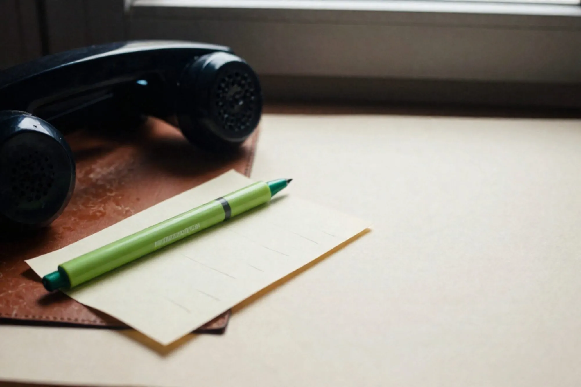 Black bakelite phone receiver on leather blotter, green silk ribbon across cord, folded cream index card beside it.
