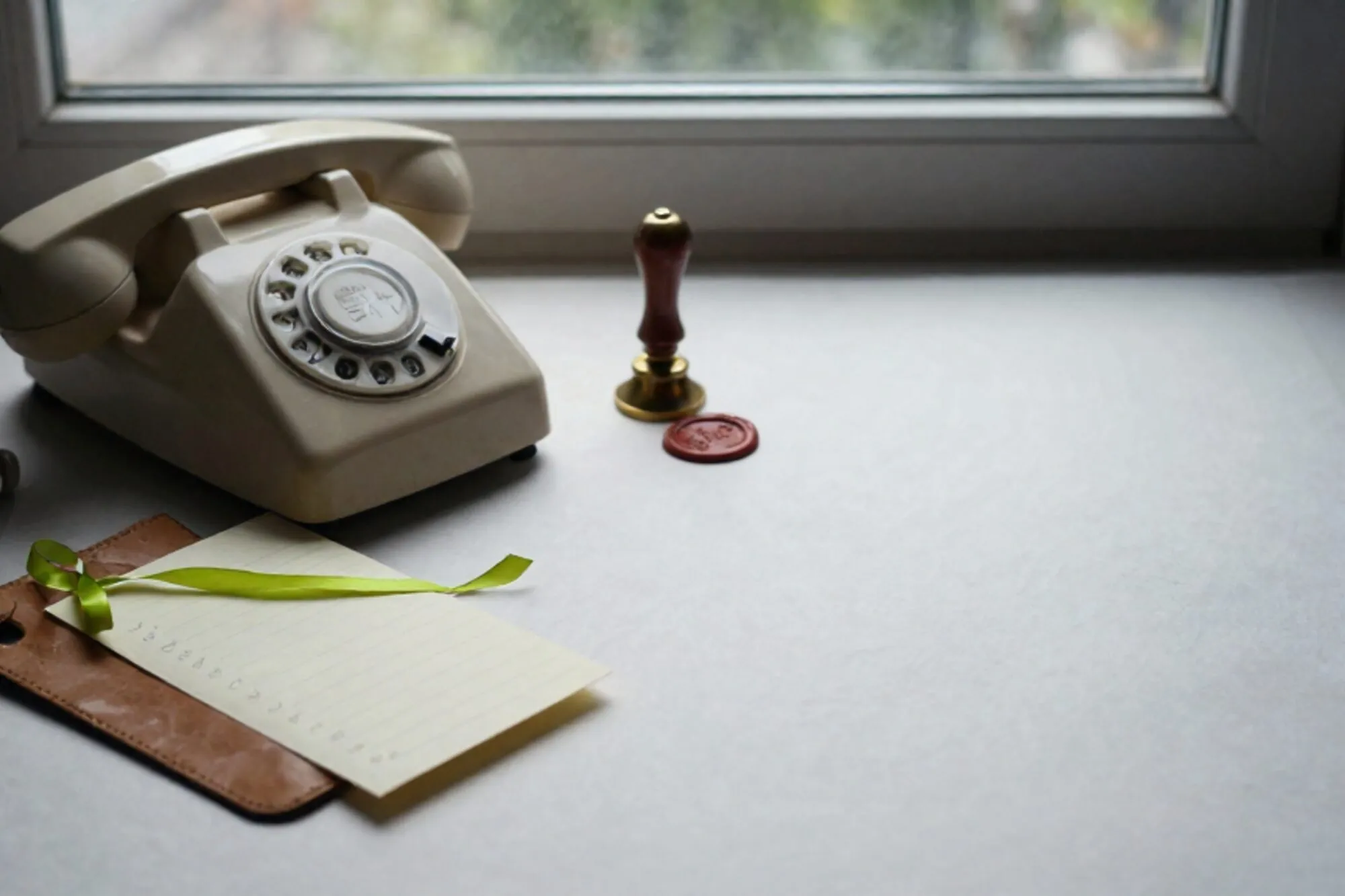 Cream bakelite phone receiver off-hook on leather blotter, green silk ribbon on index card, brass bell, wax seal.