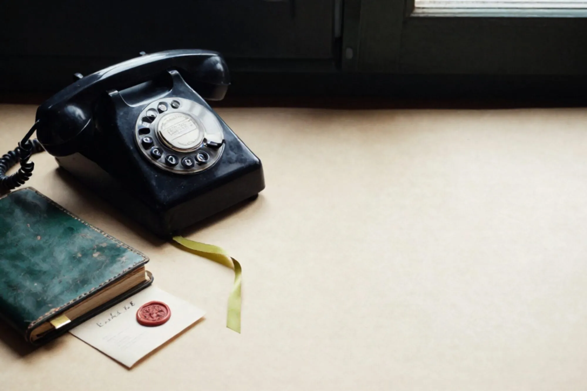 Vintage black bakelite phone receiver off-hook on forest-green leather, ledger and wax-seal card on ivory desk.