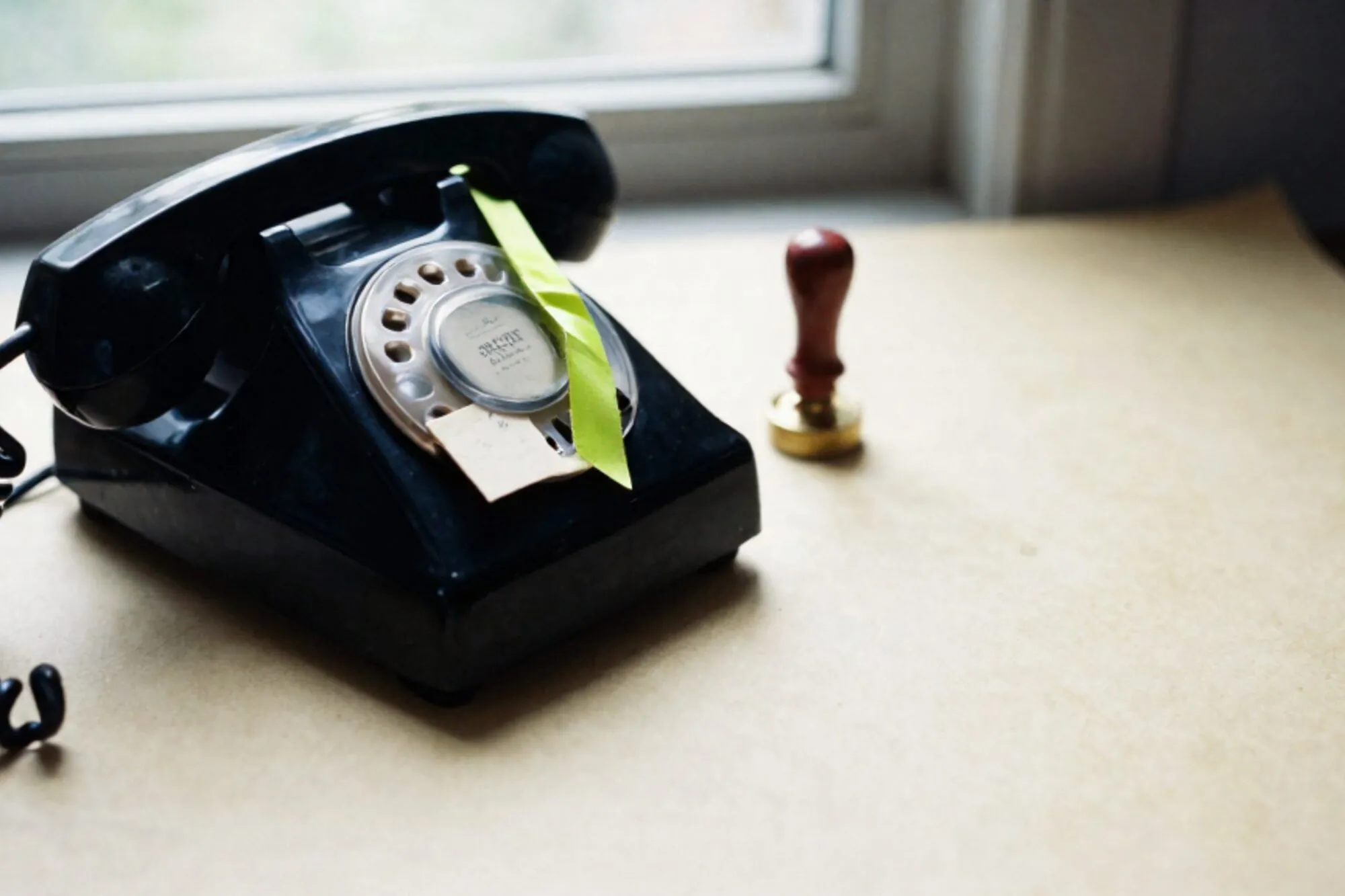 Black bakelite phone receiver off-hook on cream leather, green ribbon across it, ivory card, brass paperweight, wax seal.