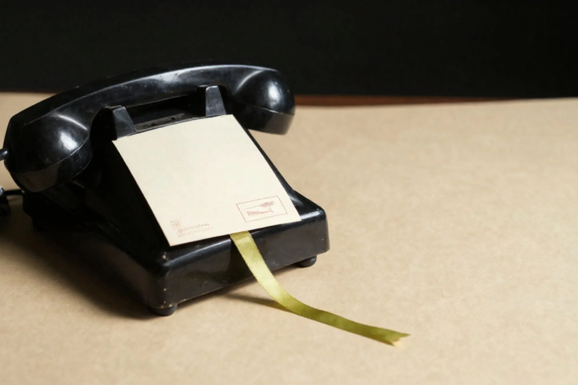 Black bakelite phone receiver off-hook on leather blotter, green ribbon, cream index card, brass plug, ivory paper.