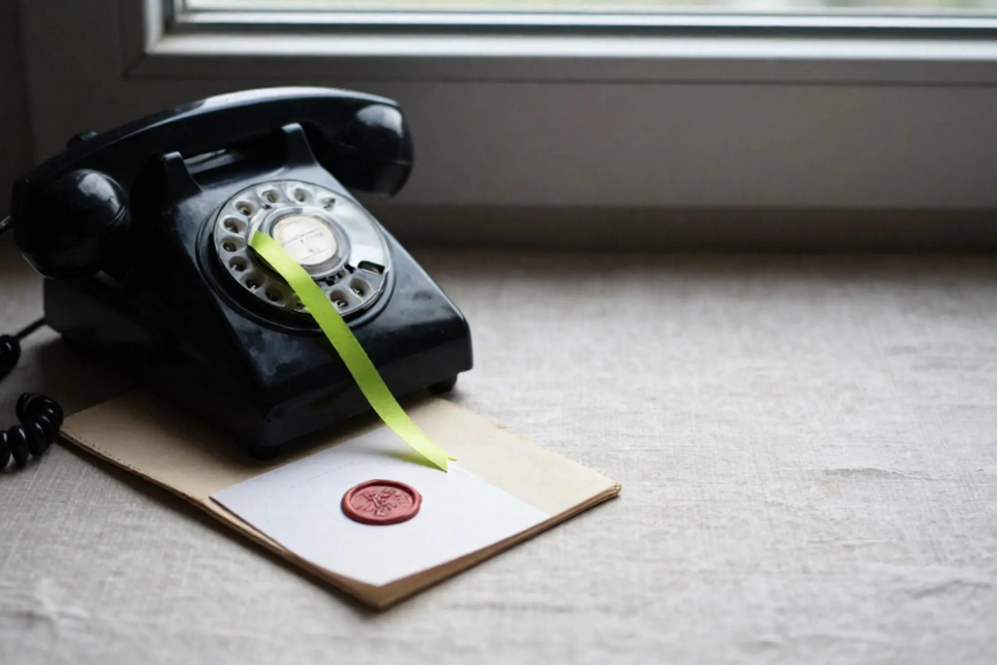 Vintage black bakelite phone receiver off-hook on cream leather blotter, green ribbon, wax-sealed card, linen surface.