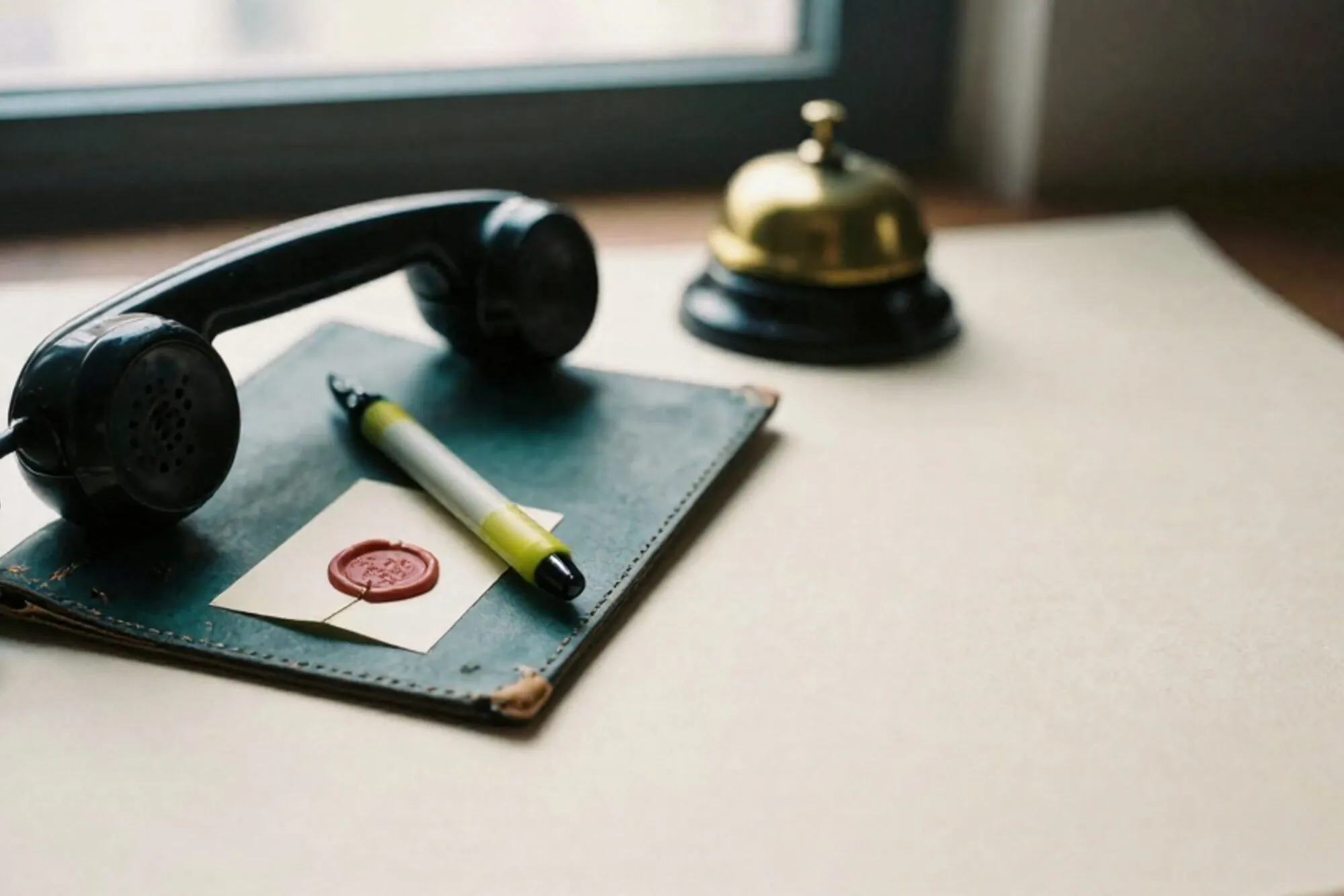 Black bakelite telephone receiver off-hook on green leather blotter, green ribbon, wax-sealed paper, brass bell behind.