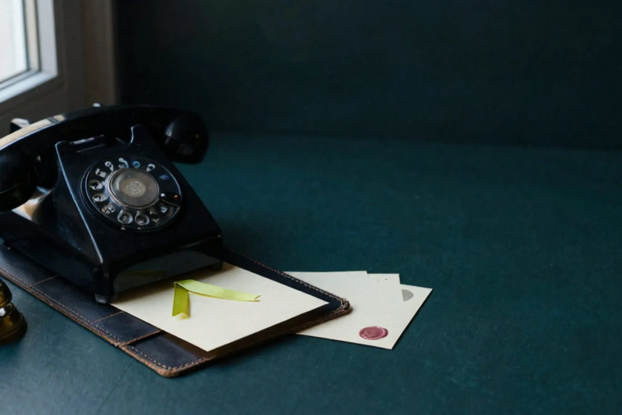 Black bakelite phone receiver off-hook on leather blotter, paper note with green ribbon, ivory cards, brass bell.