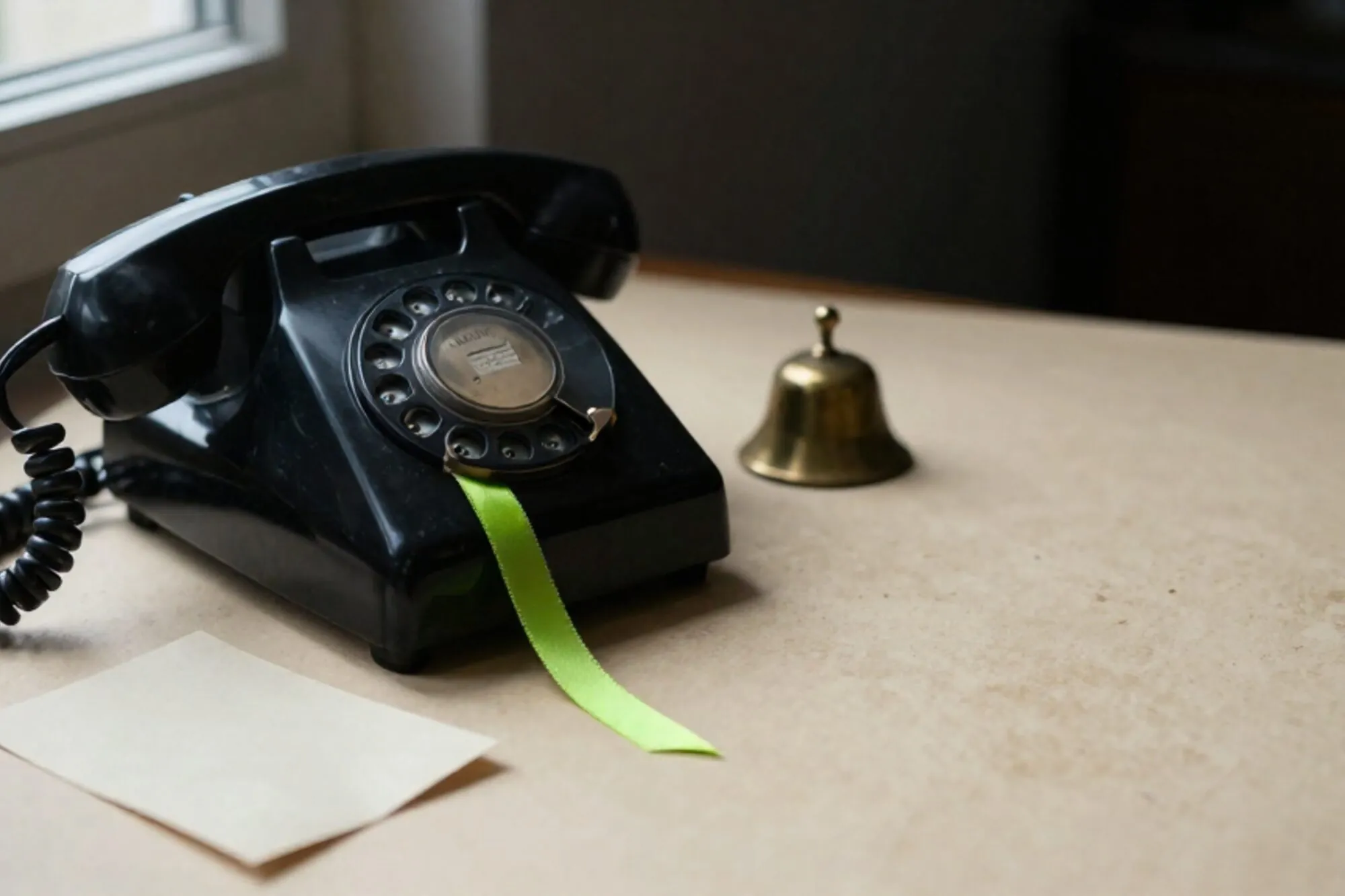 Zwarte bakelieten telefoonhoorn op leren onderlegger, gekrulde snoer, groen lint, messing belletje, ivoren kaart.