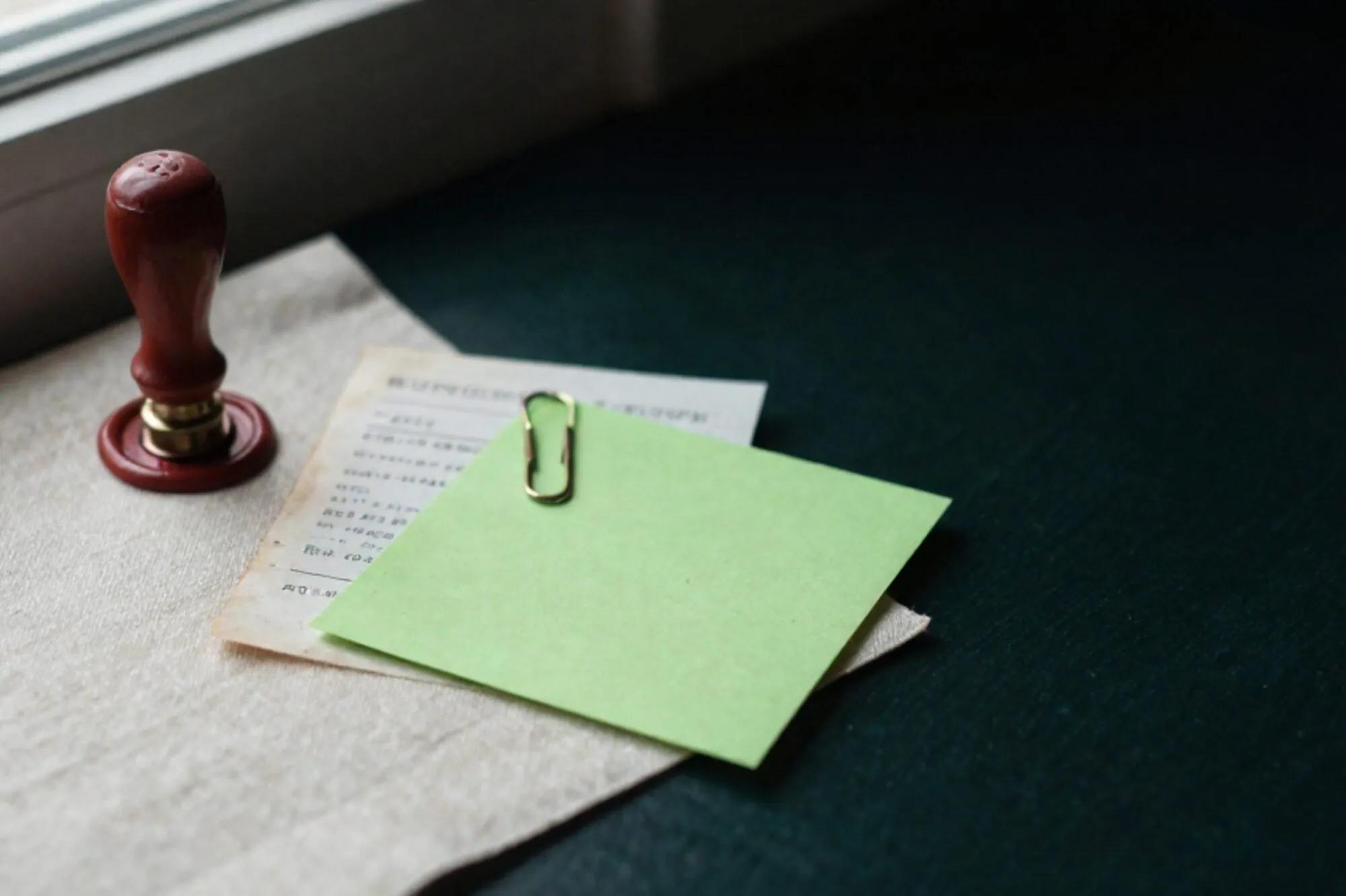 Worn appointment card on ivory linen, brass paperclip with green sticky note, carbon receipt, red wax seal, dark shadow.