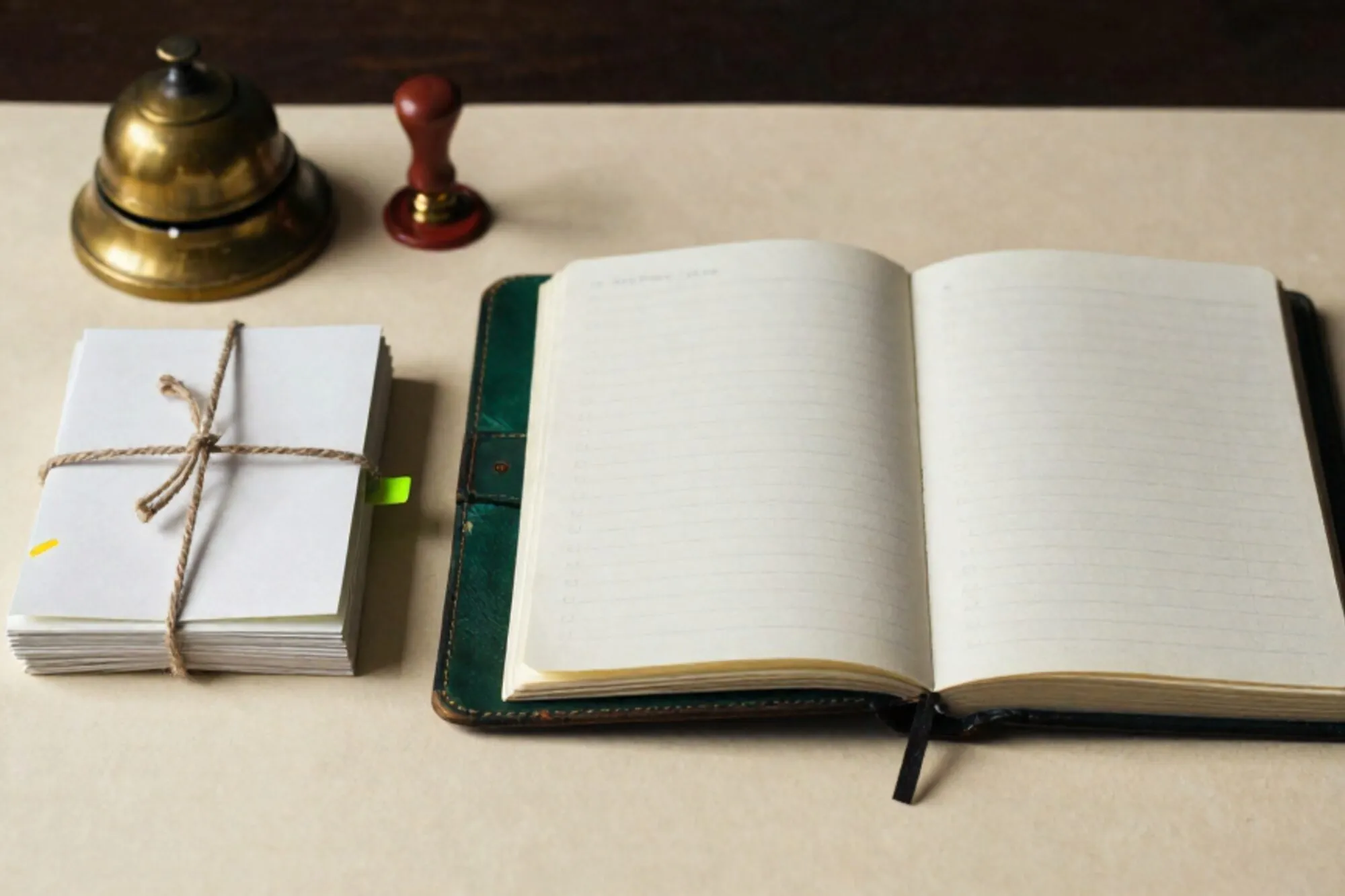 Open leather logbook on ivory desk, paper slips tied with twine, green tab, brass bell, red wax seal, side light.