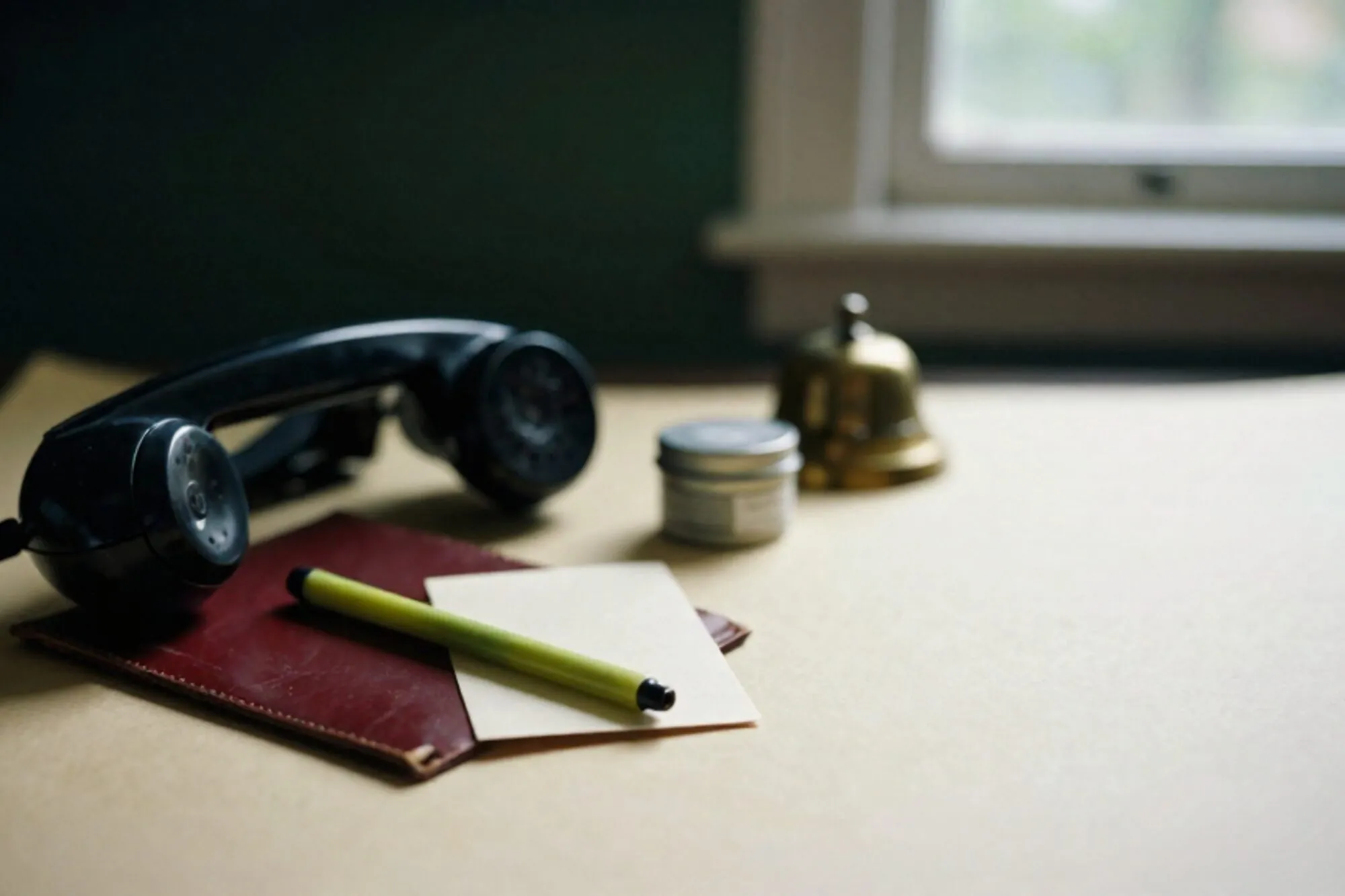 Black bakelite phone receiver off-hook on oxblood leather blotter, green ribbon on cream card, brass bell behind.