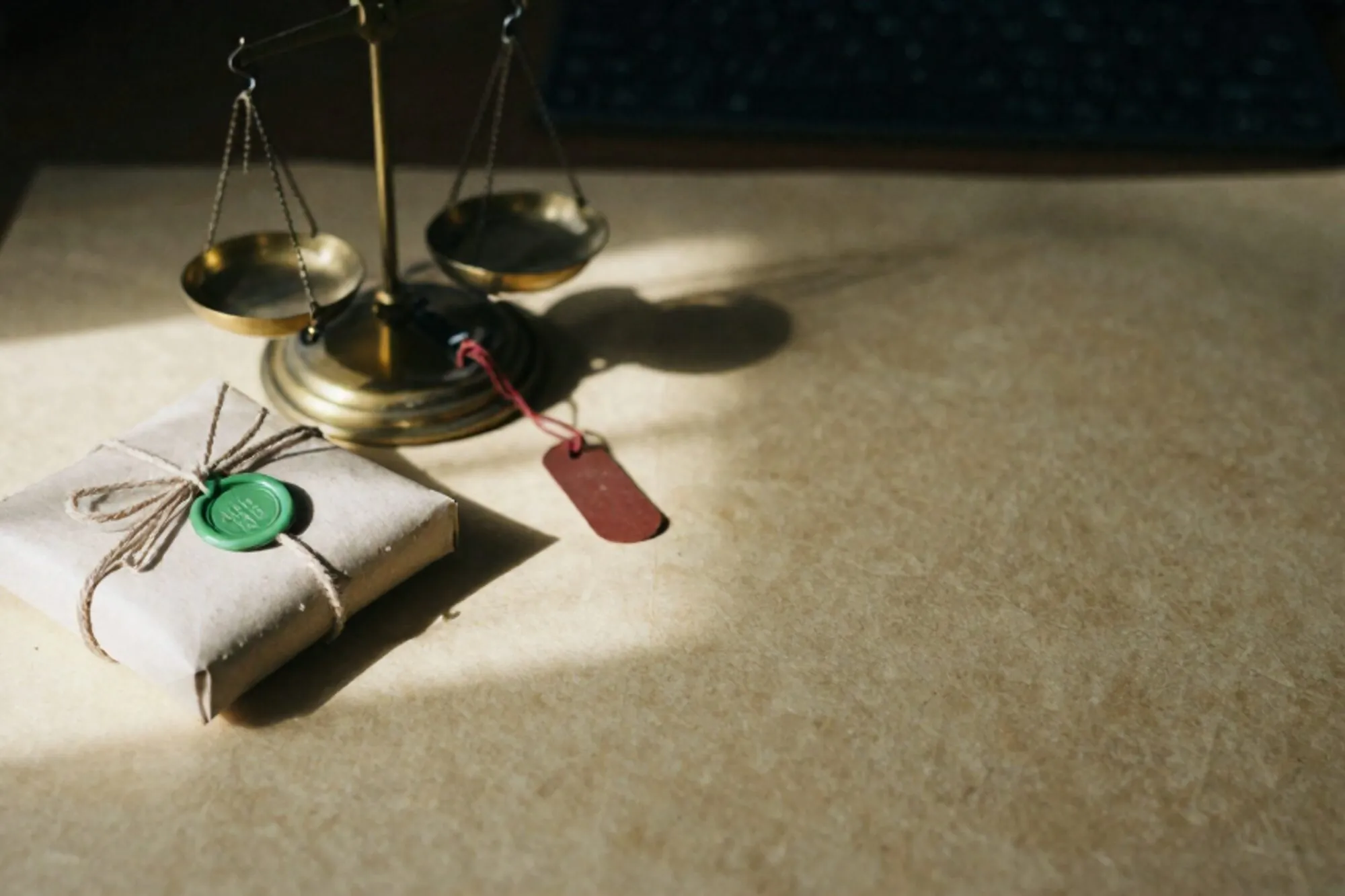 Craft-paper parcel with green wax seal beside a tilted brass scale with red tag on an ivory desk.