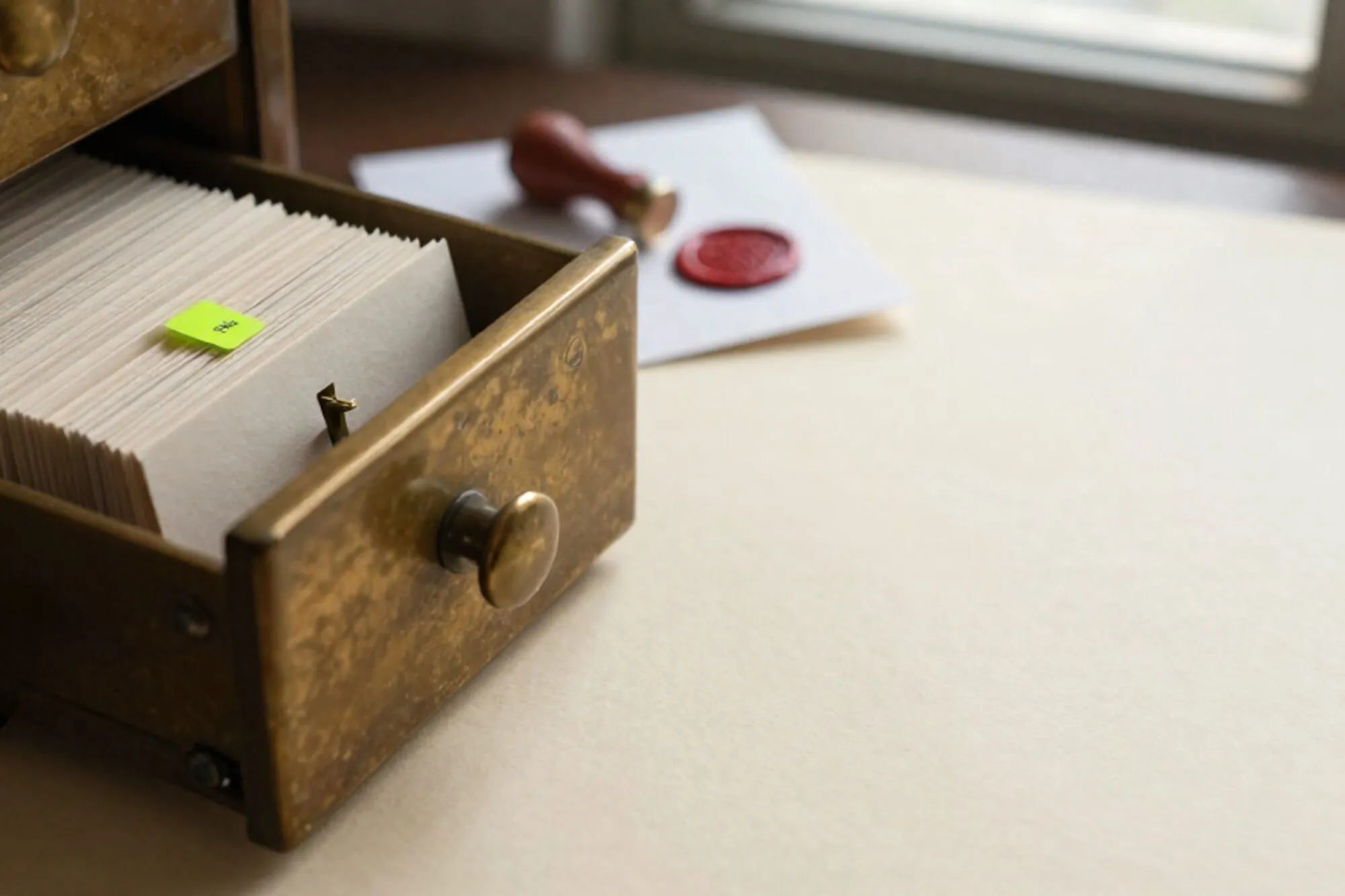 Open brass card-catalog drawer with index cards, one chartreuse tab raised, wax seal on folded slip, ivory paper surface.