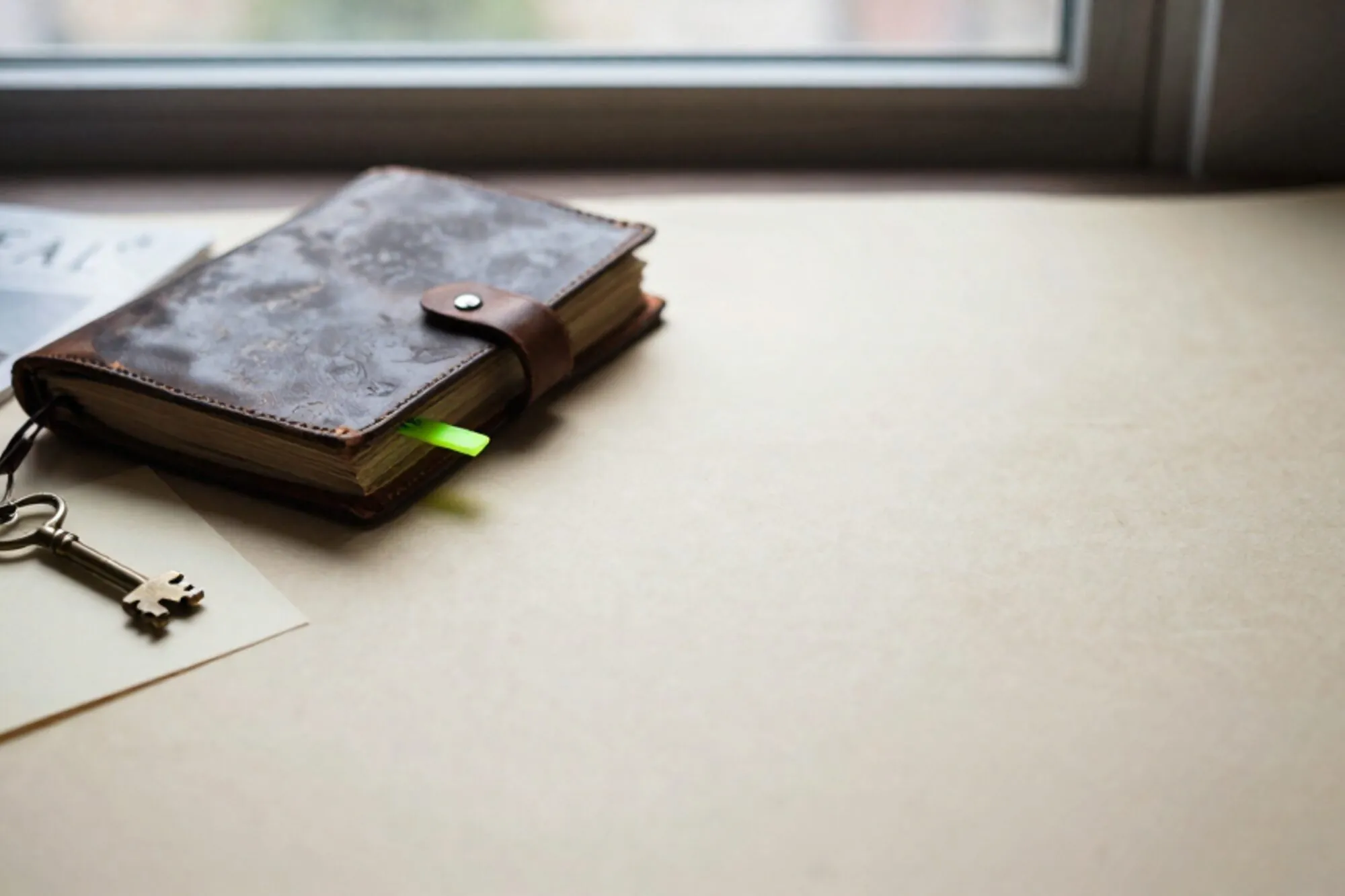 Open leather logbook, brass key on cream card, green ribbon marker on ivory paper, side window light.