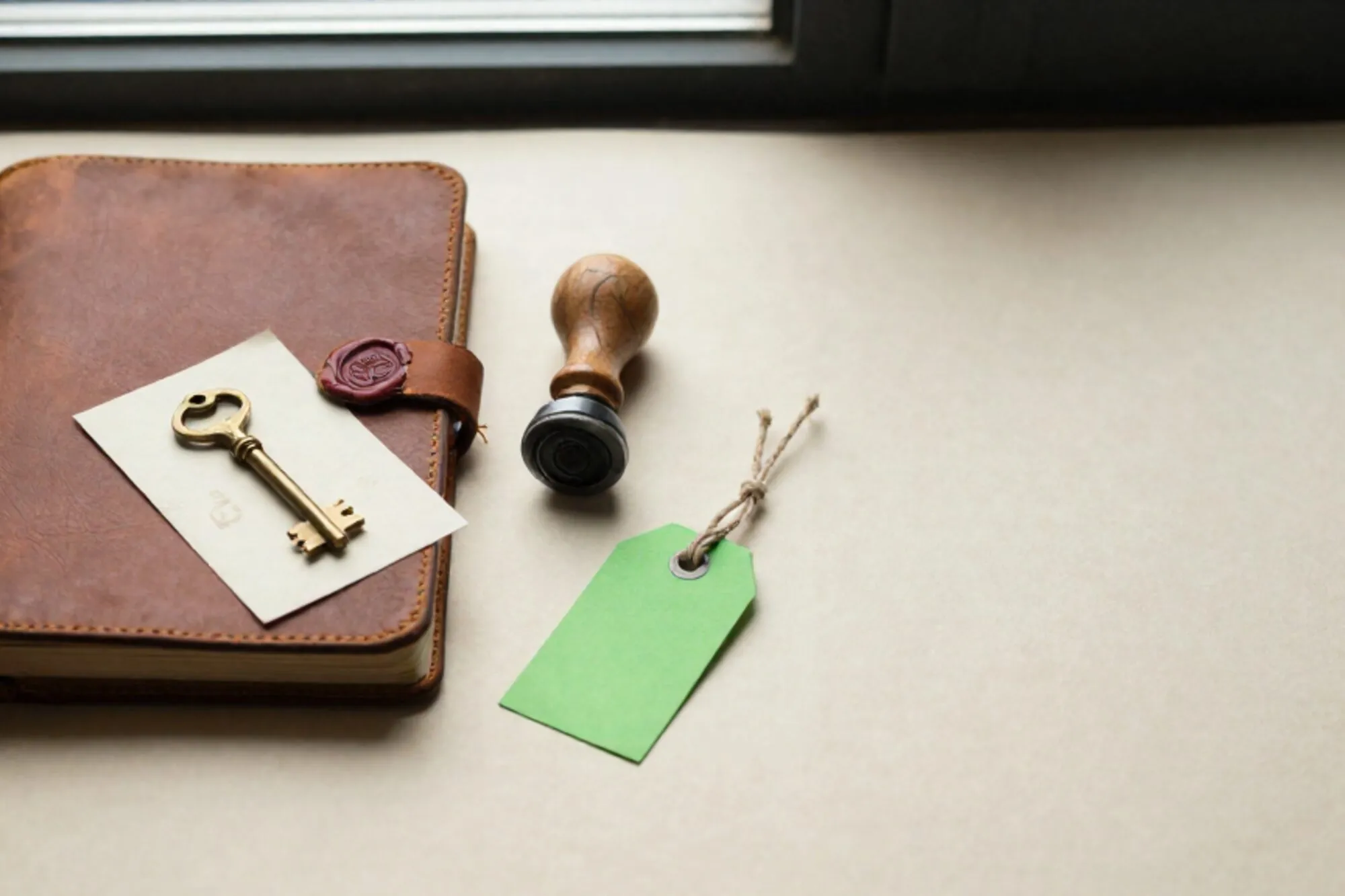 Closed leather logbook with silk ribbon, brass key on index card, rubber stamp, green tag on ivory paper.