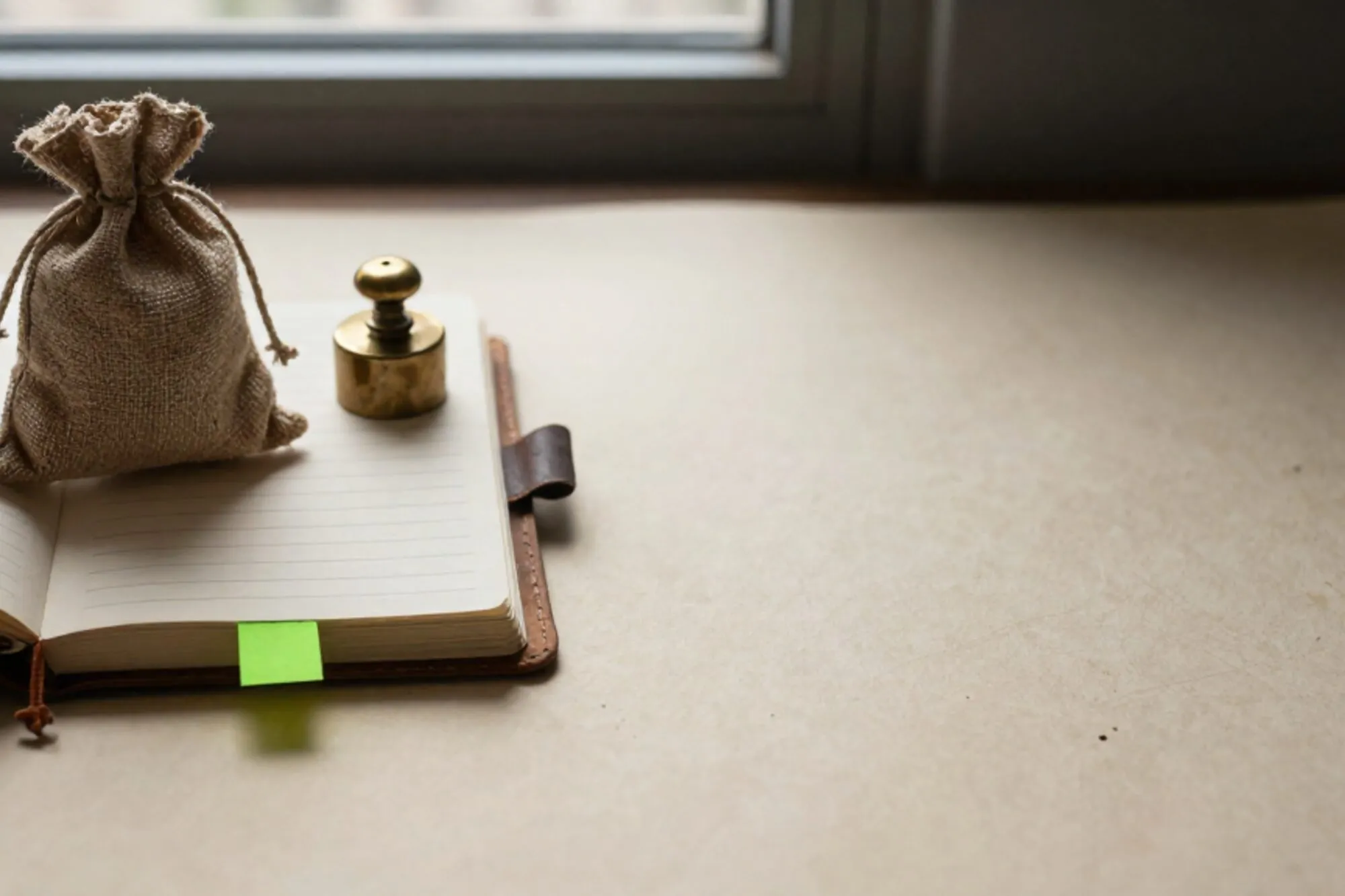 Leather ledger half-open on ivory paper, burlap coffee sack with twine, brass weight, green bookmark, red wax seal.