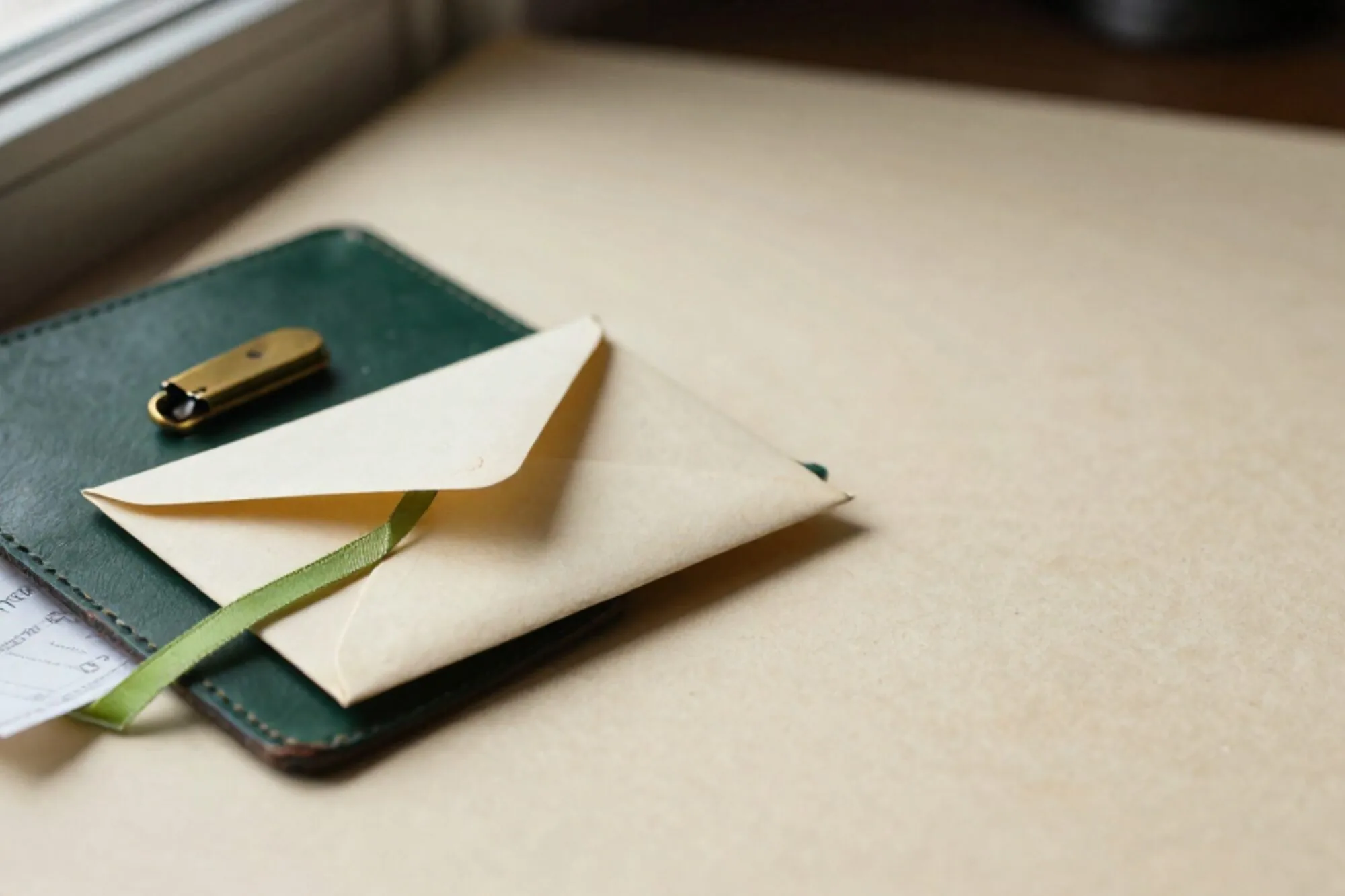 Crèmekleurige envelop met limegroen lint op donkergroen leren vloeiblad, koperen briefopener en gevouwen bonnetje ernaast.